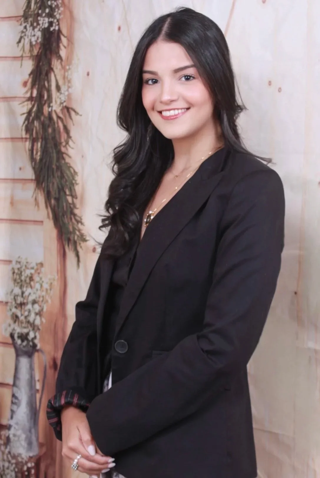 A woman with long dark hair wearing a black blazer and smiling, standing indoors with a wooden wall and dried plants in the background.