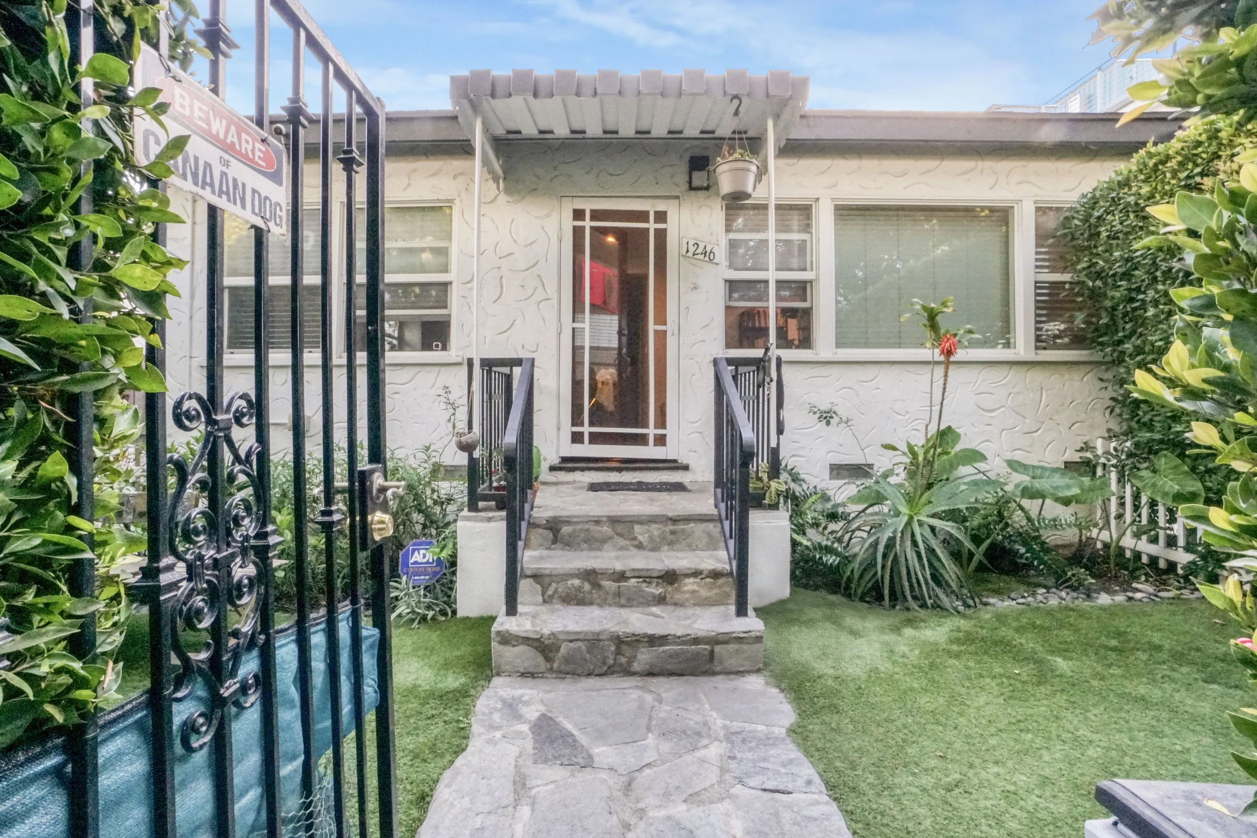 Front entrance of a house with stone steps, black metal railing, surrounded by green bushes and plants, with a white house exterior and windows, a small covered porch, and a gate with a