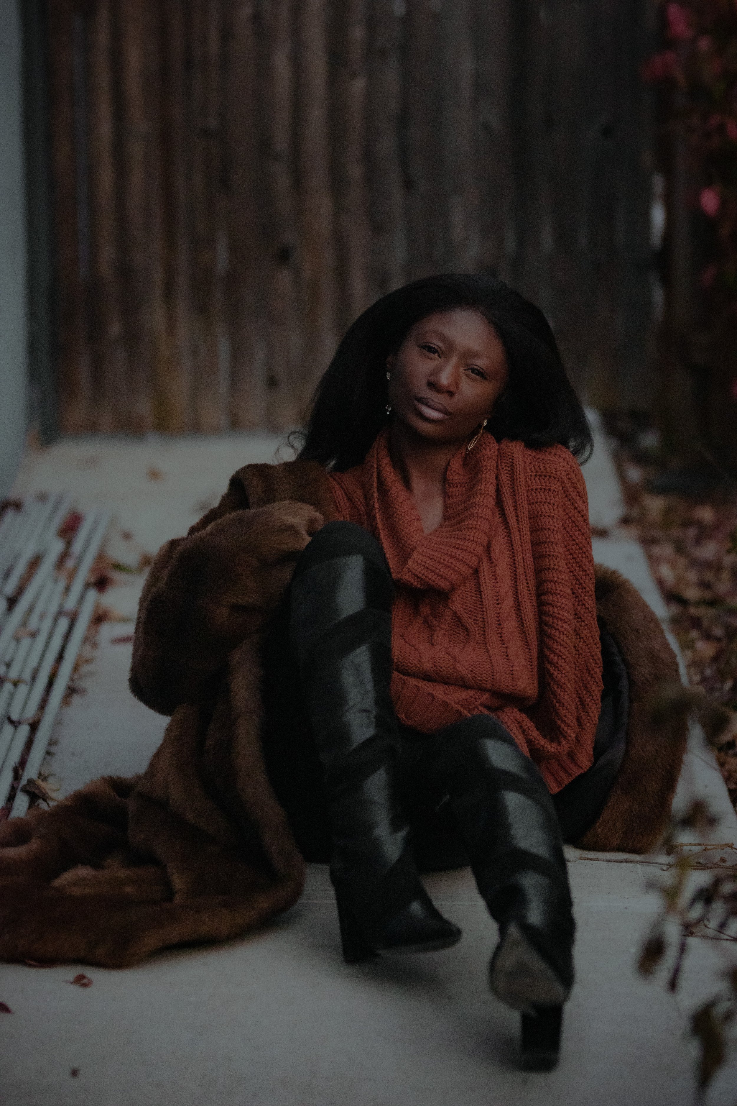 Picture of Joy Ayoka seated on palish grey concrete wearing a burnt sienna colored, thick-knit cowl neck sweater and black leggings/knee high boots. She wears a near floor length Chocolate Brown Faux Fur Coat.