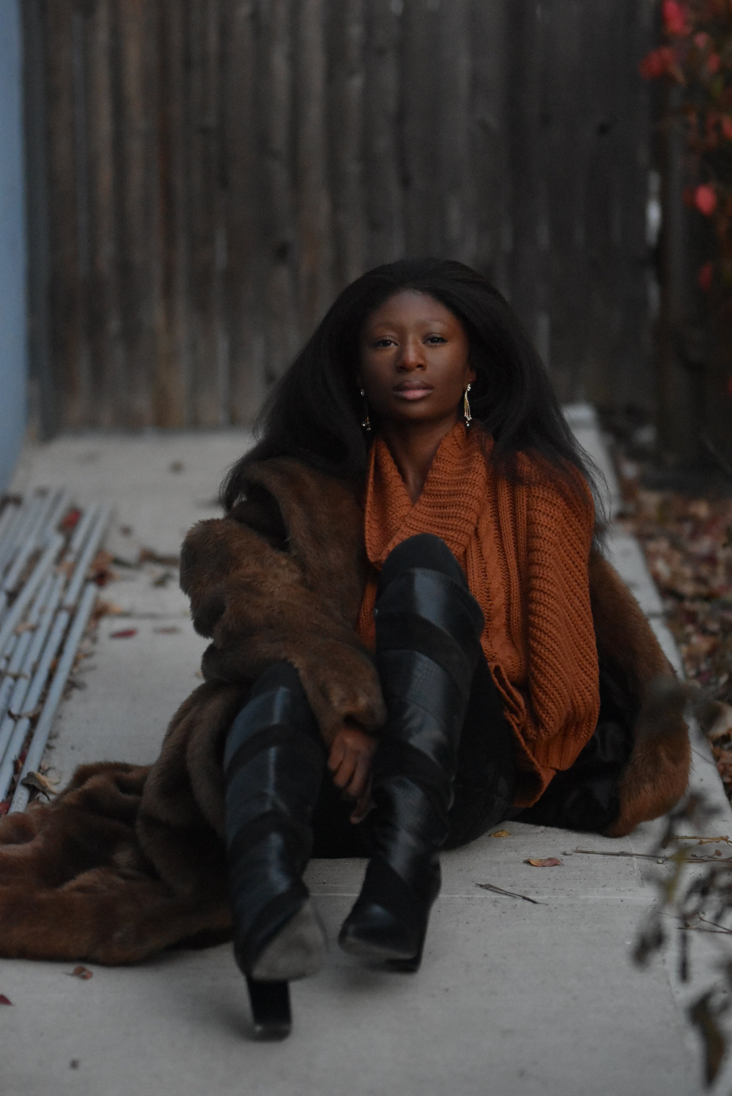 Picture of Joy Ayoka seated on palish grey concrete wearing a burnt sienna colored, thick-knit cowl neck sweater and black leggings/knee high boots. She wears a near floor length Chocolate Brown Faux Fur Coat.