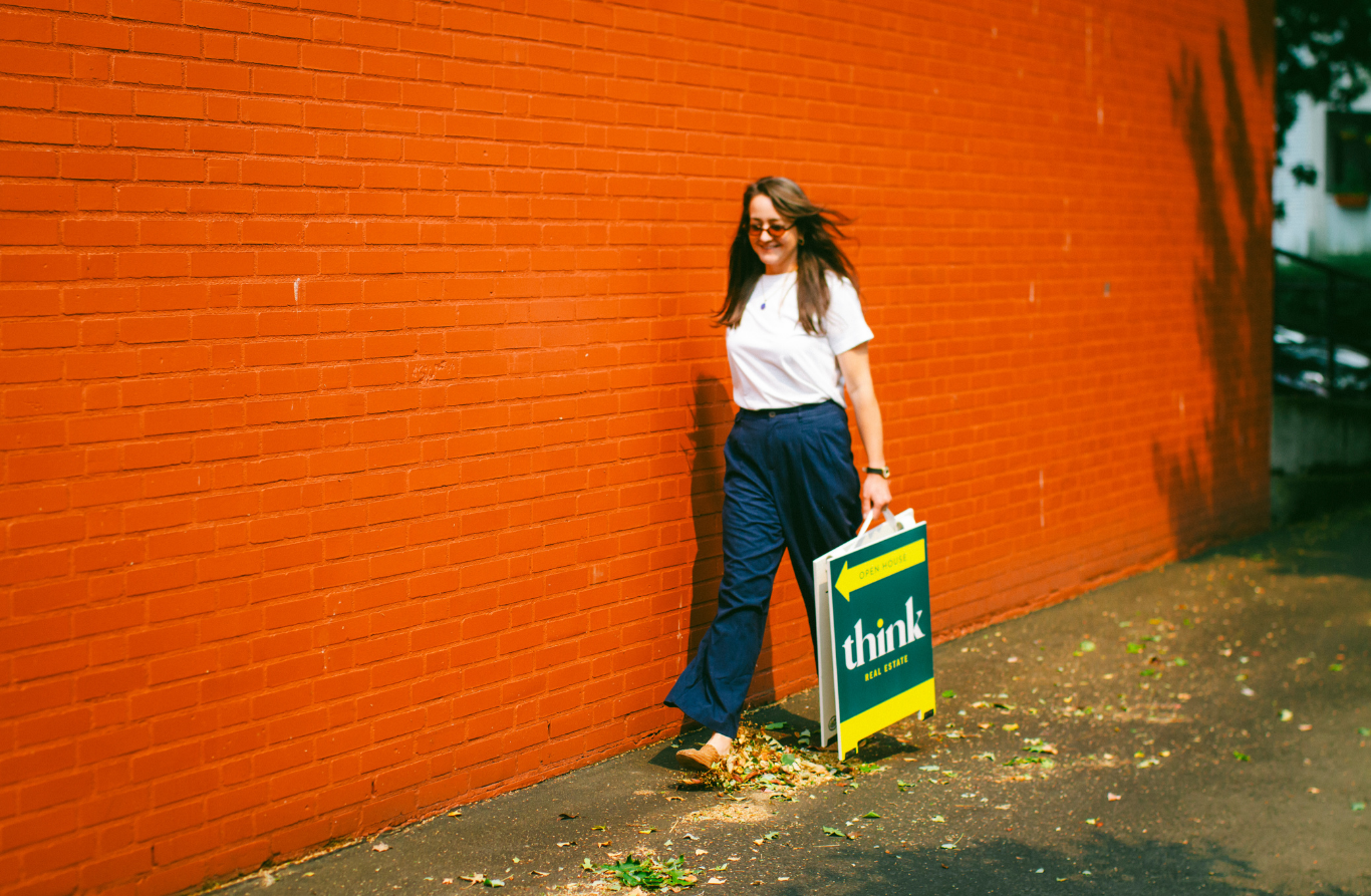 Woman standing against a red brick wall holding a real estate sign that reads "think REAL ESTATE" and has an arrow pointing left.