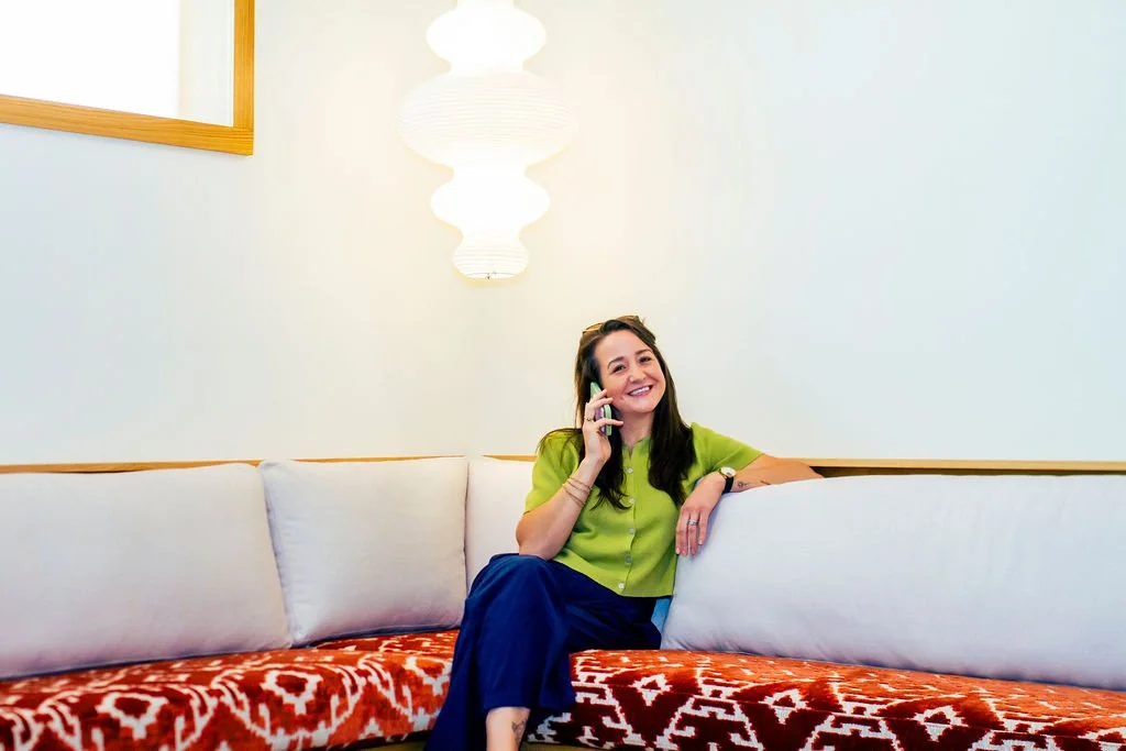 A woman smiling and talking on her cellphone while sitting on a sofa in a brightly lit room.