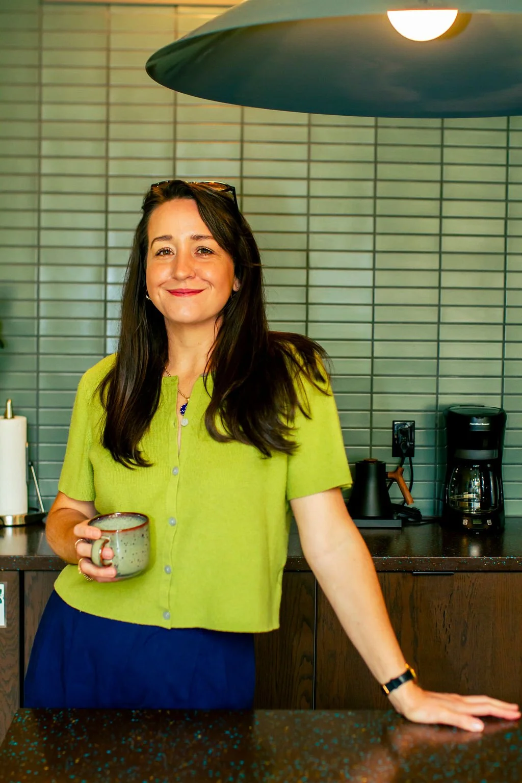 A woman with long dark hair, wearing a lime green top and a dark bracelet, standing behind a dark speckled countertop in a kitchen, holding a ceramic mug and smiling at the camera.