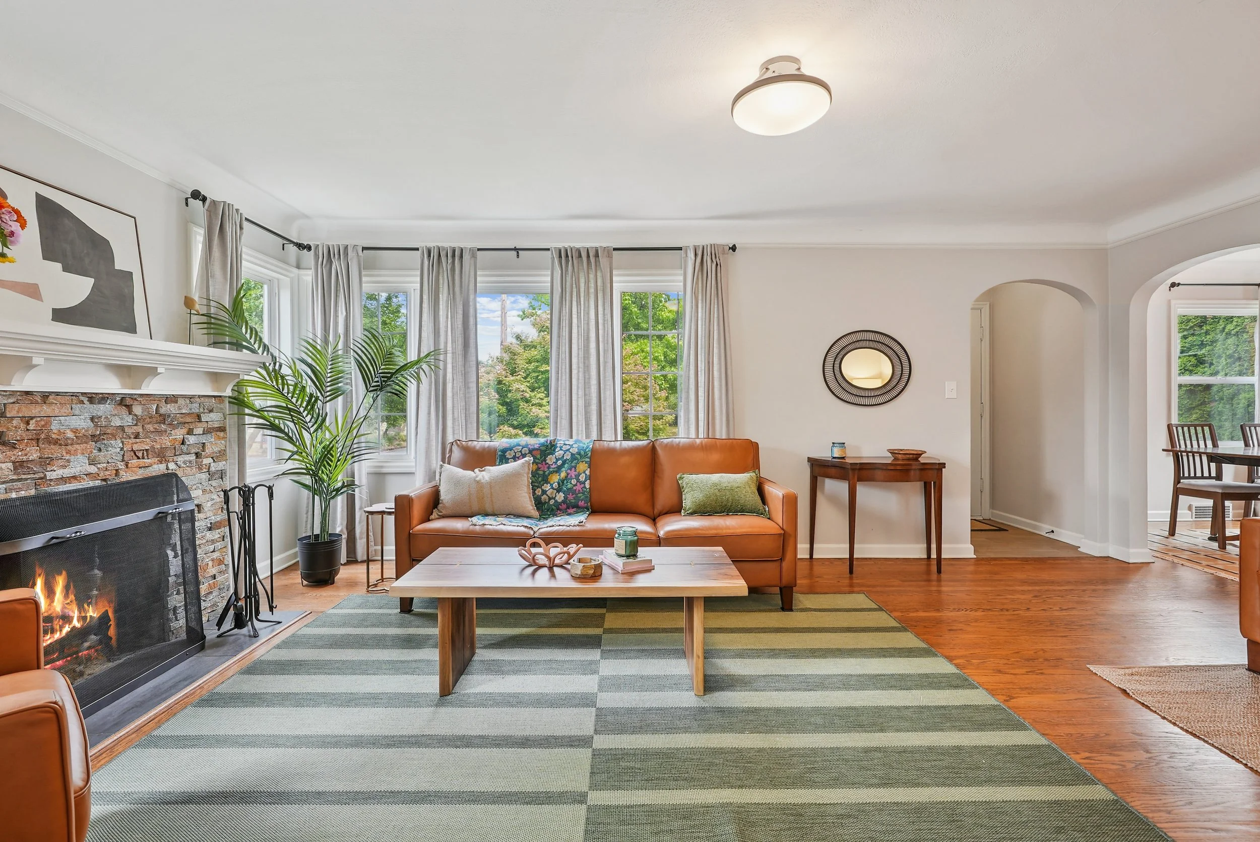 Living room with large windows, beige curtains, a brick fireplace, a leather sofa with pillows, a wooden coffee table, and indoor plants.
