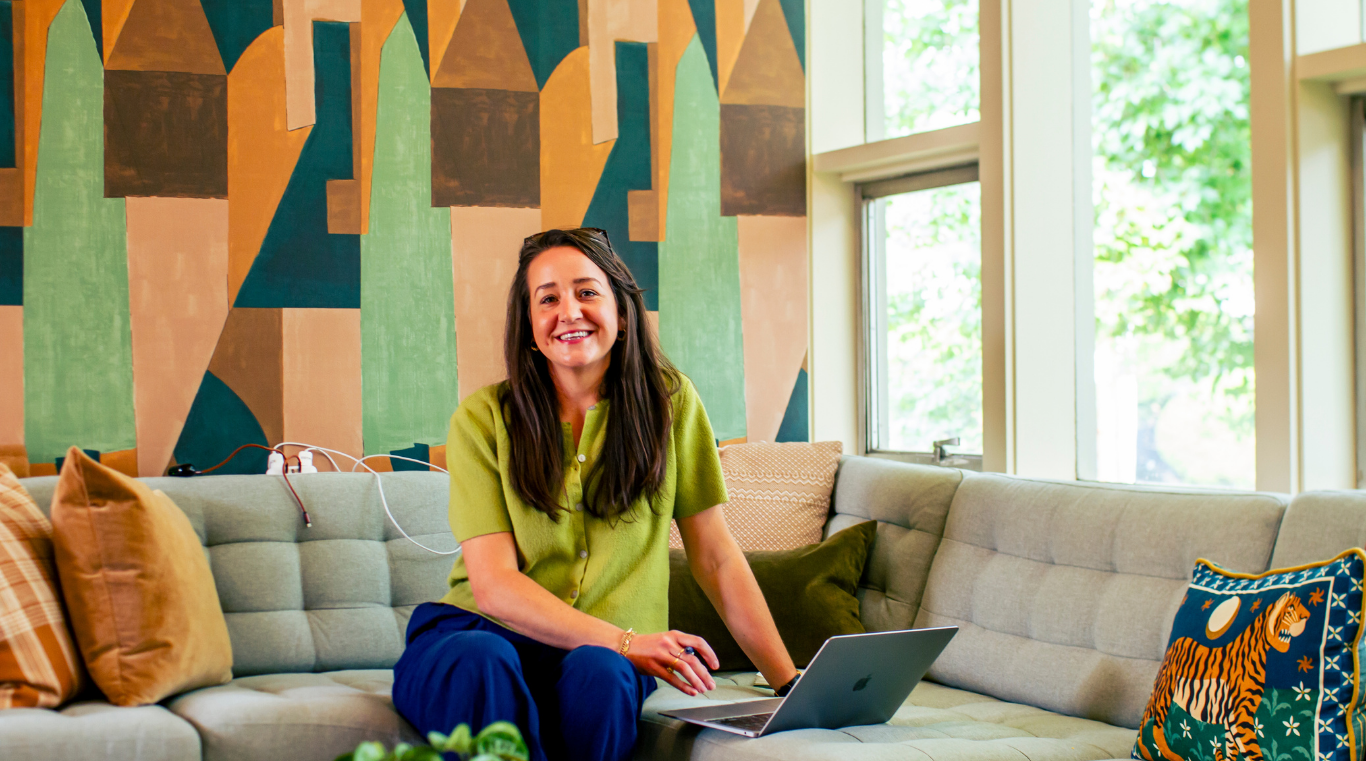 A woman with long dark hair wearing a bright green shirt and blue pants is sitting on a beige sofa in a room with large windows. She is smiling and working on a silver laptop. The room has colorful pillows, a patterned wall behind her, and lush green trees visible through the windows.
