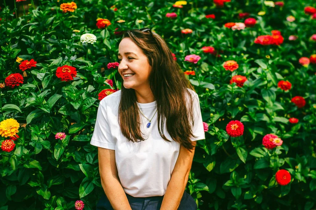 A woman with long brown hair smiling and wearing a white t-shirt, sitting in a vibrant flower garden filled with red, pink, yellow, and white flowers.