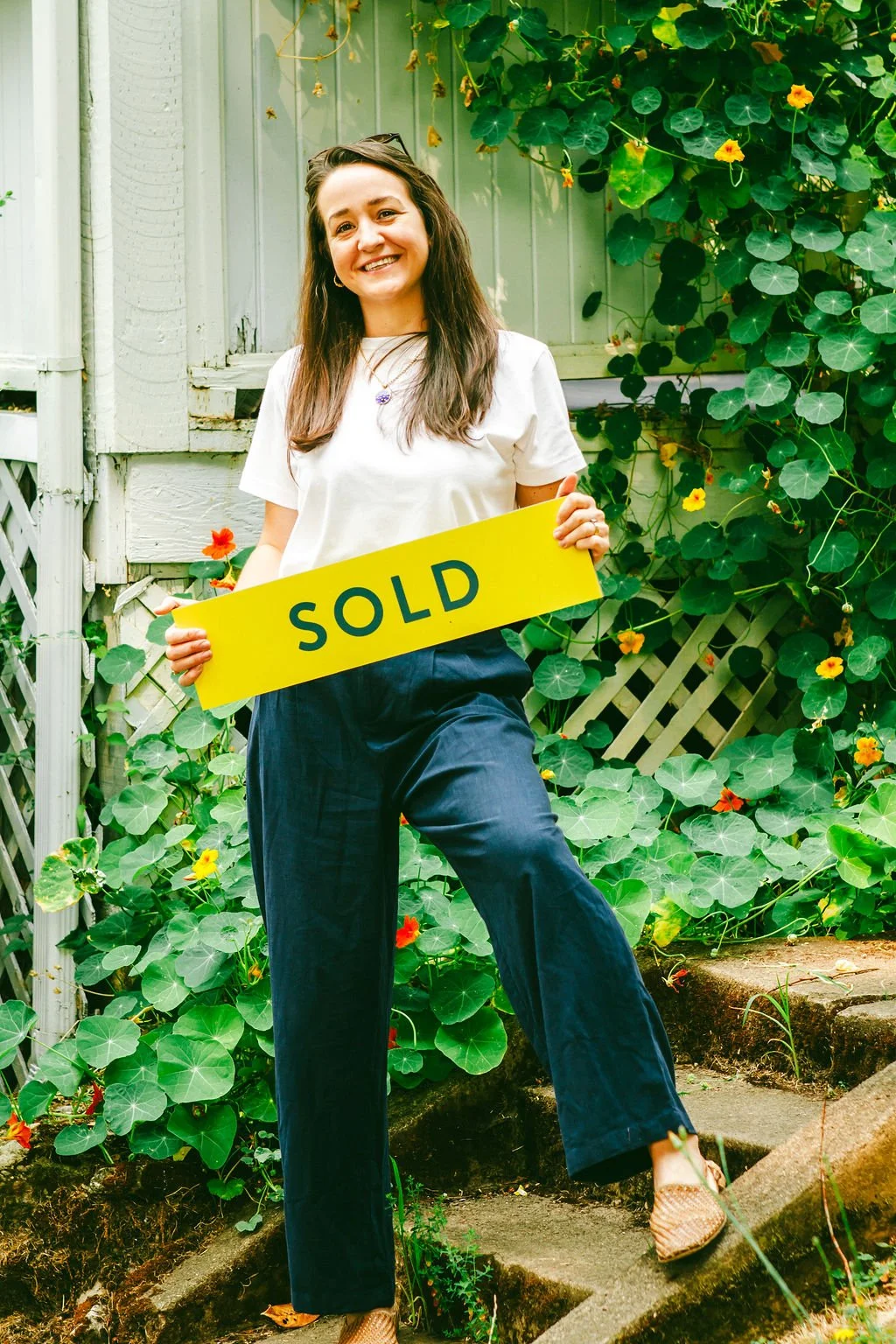 A woman smiling and holding a yellow sign that says 'SOLD' standing outdoors in front of a garden with green foliage and flowers.