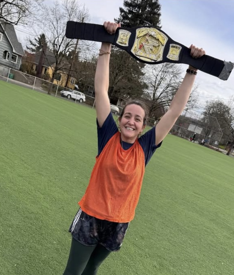 A woman standing on a soccer field, holding a championship belt above her head, smiling.