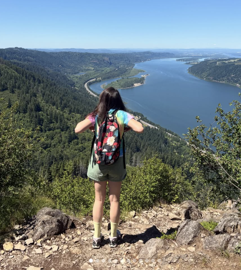 A girl with long dark hair wearing a colorful tie-dye shirt, shorts, and hiking shoes, standing on a rocky trail with a backpack, overlooking a large winding river surrounded by green forested hills under a clear blue sky.