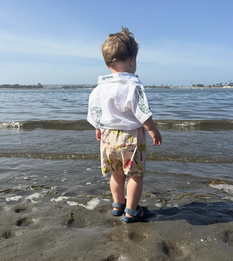 A young boy standing on a sandy beach near the water, wearing a white shirt, patterned shorts, and sandals, looking out at the ocean with a clear sky overhead.