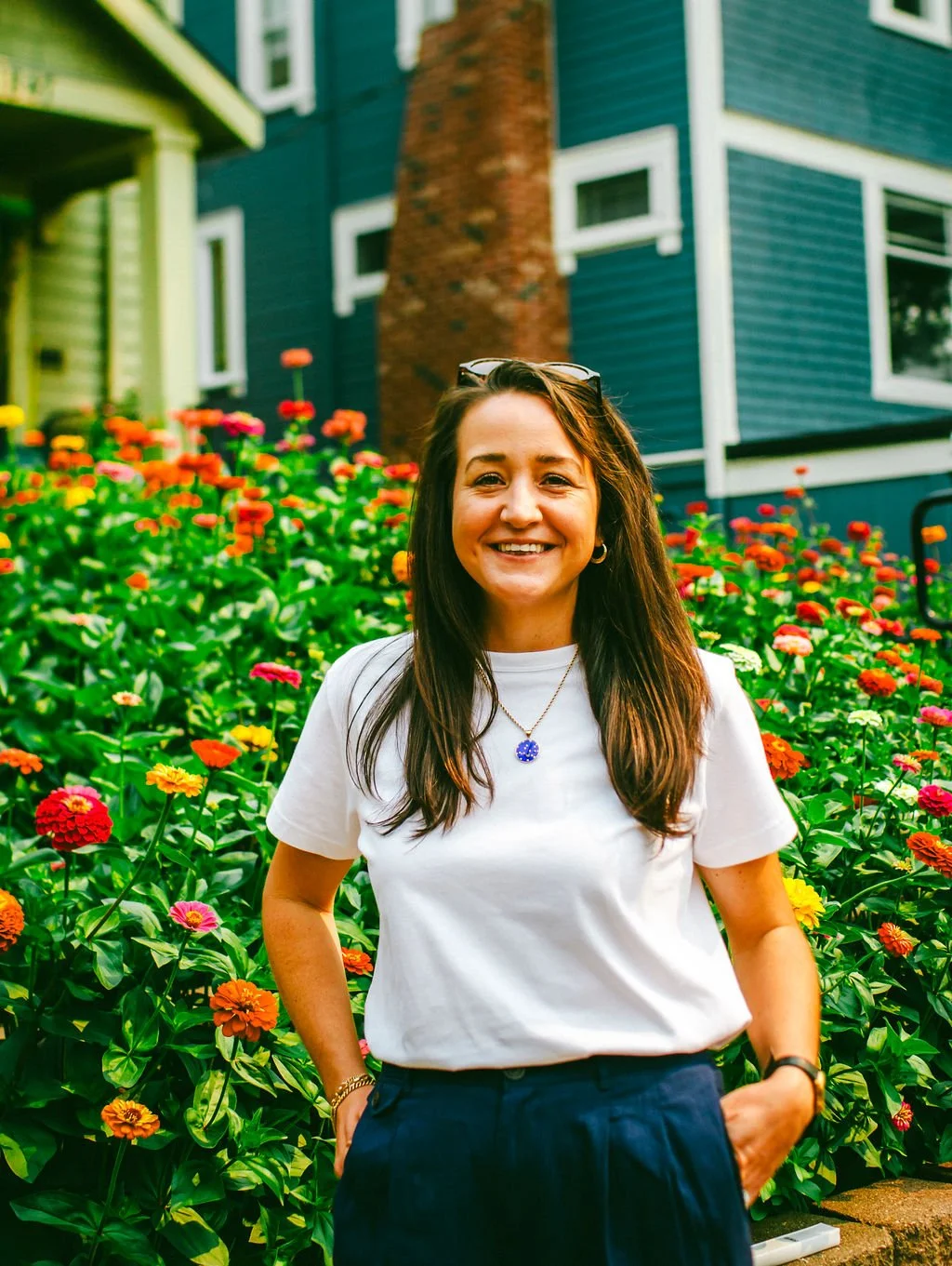 A woman with long brown hair smiling, wearing a white t-shirt and dark pants, standing in front of a colorful flower garden with a house in the background.