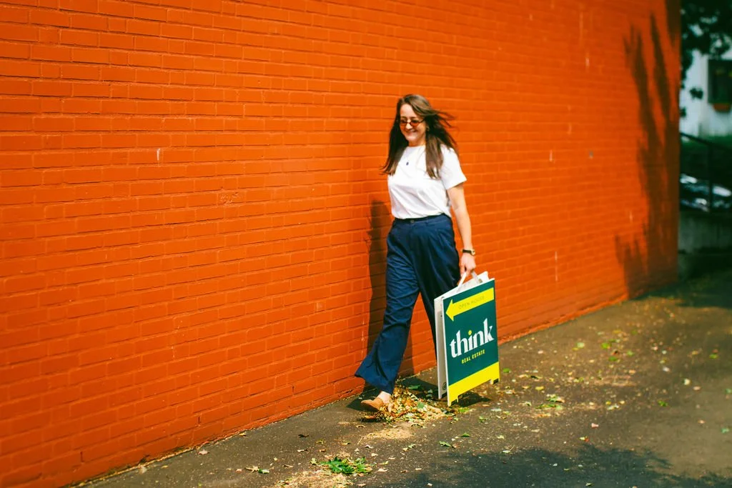 Woman with long hair, wearing glasses, a white shirt, and navy pants, standing next to a sidewalk sign that reads 'think' with an arrow pointing left against a red brick wall.