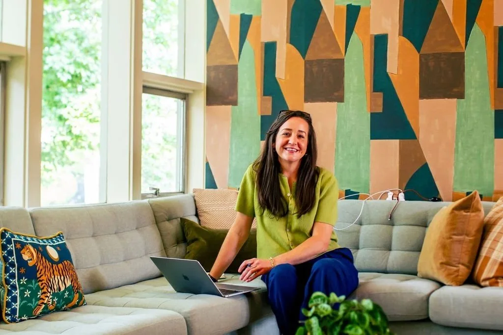 A woman sitting on a light gray sectional sofa in a well-lit room with large windows, colorful geometric wall art, and decorative pillows, working on a laptop.