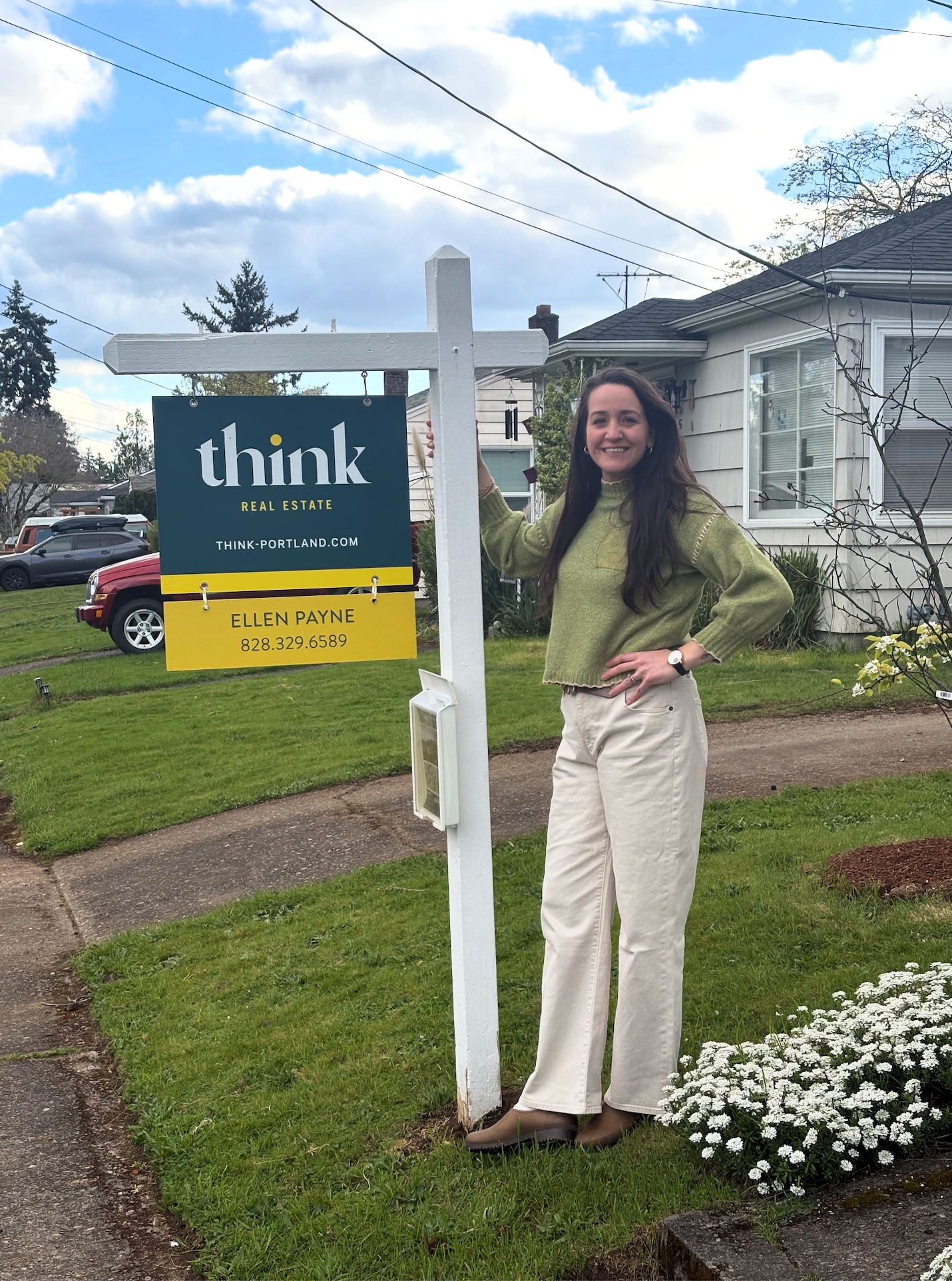 A woman standing next to a real estate sign on a residential lawn, smiling at the camera. She has long, dark hair and is wearing a green sweater, white pants, and brown boots. The sign displays the name Ellen Payne, the website think-portland.com, and a phone number.