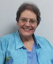 Lorraine Stoltzfus, a smiling woman with glasses and brown hair, wearing a light blue blouse, stands in front of a white wall.