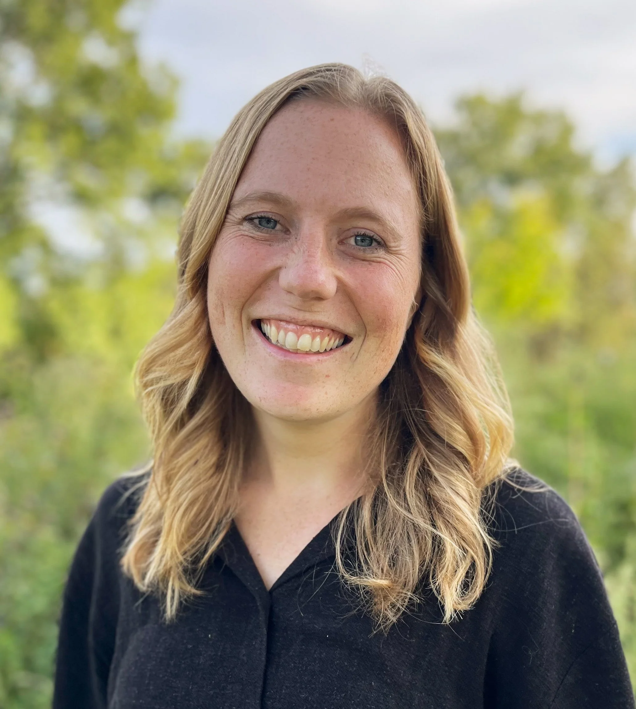 Anika Reynar, a woman with blonde hair and a black shirt, smiles in front of a blurred tree background.