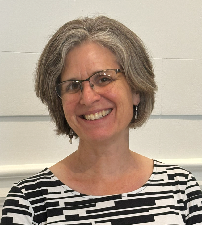Kirstin De Mello, a smiling woman with glasses and short gray hair, wearing a black and white shirt, stands in front of a white wall.