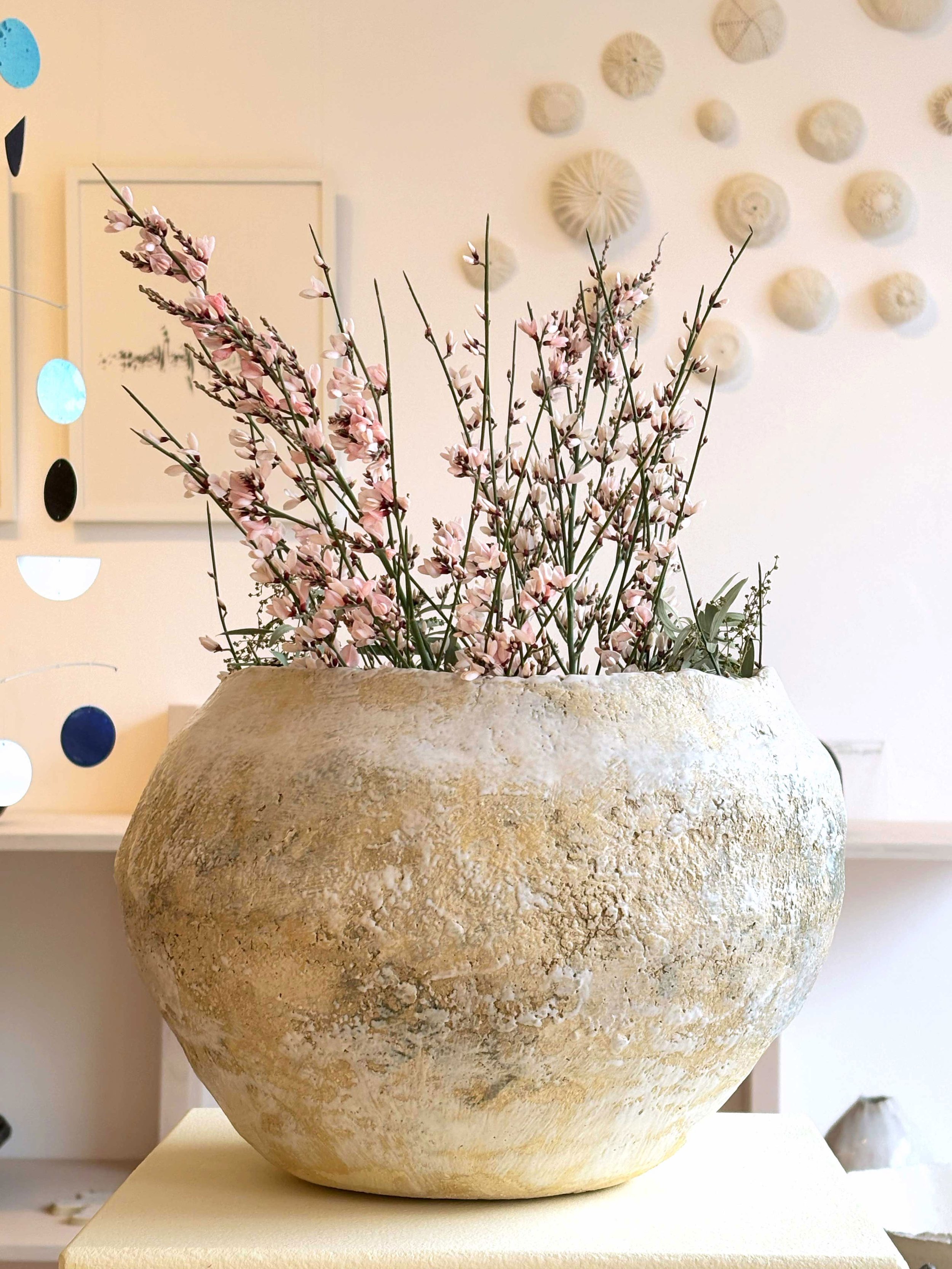 Große handgebaute Keramikvase mit rose Blumen, auf hellem Podest, im Hintergrund weiße Wand mit weissen Wandobjekten