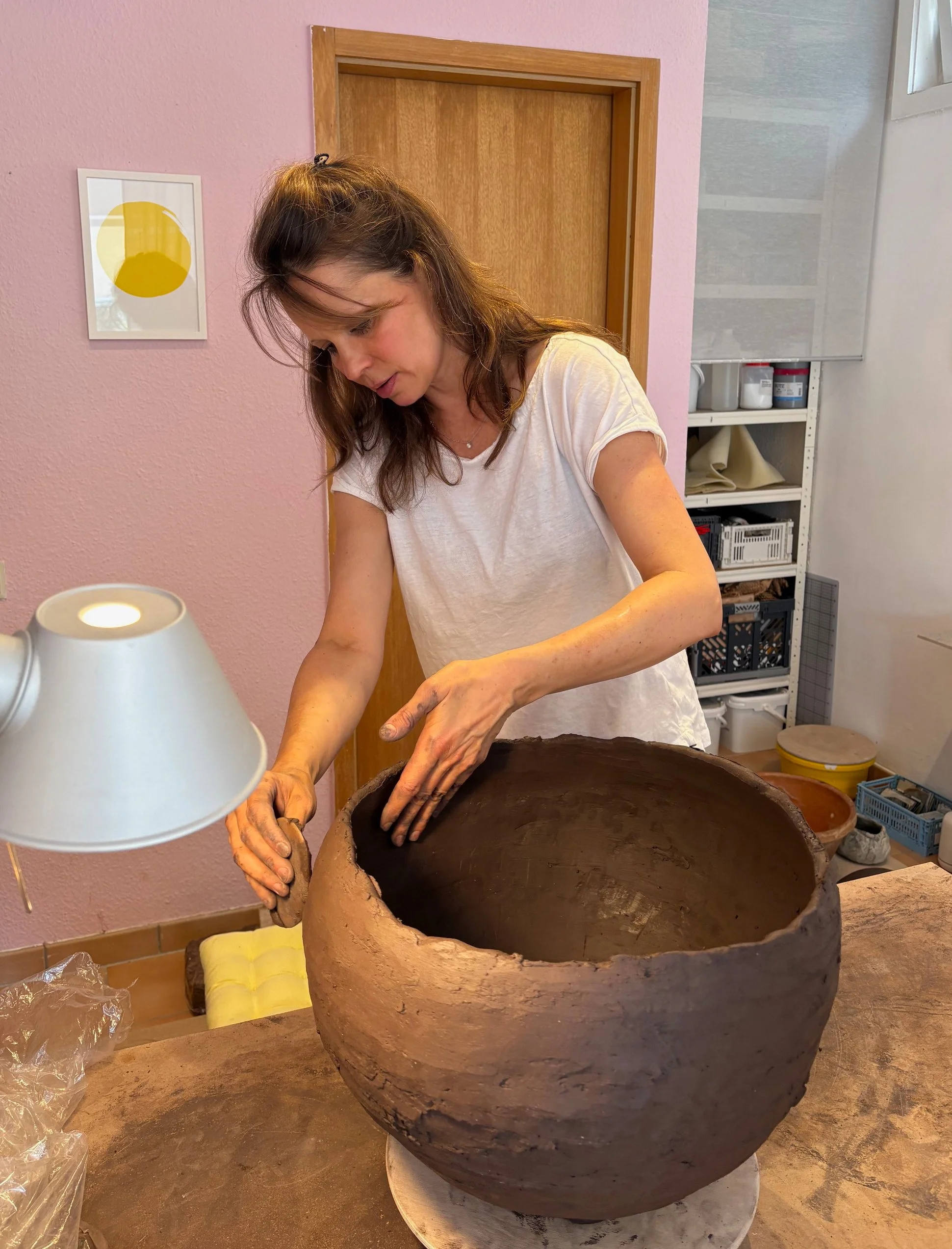 Eine Frau arbeitet an einem großen, runden Keramikgefäss aus Ton. Im Hintergrund sind Regale mit Kunstmaterialien.