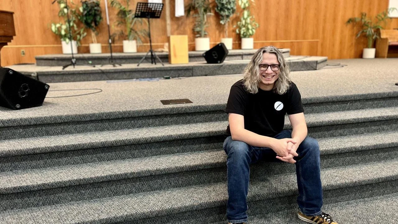 Pastor James Sholl sits on the stairs of the sanctuary stage and smiles at the camera