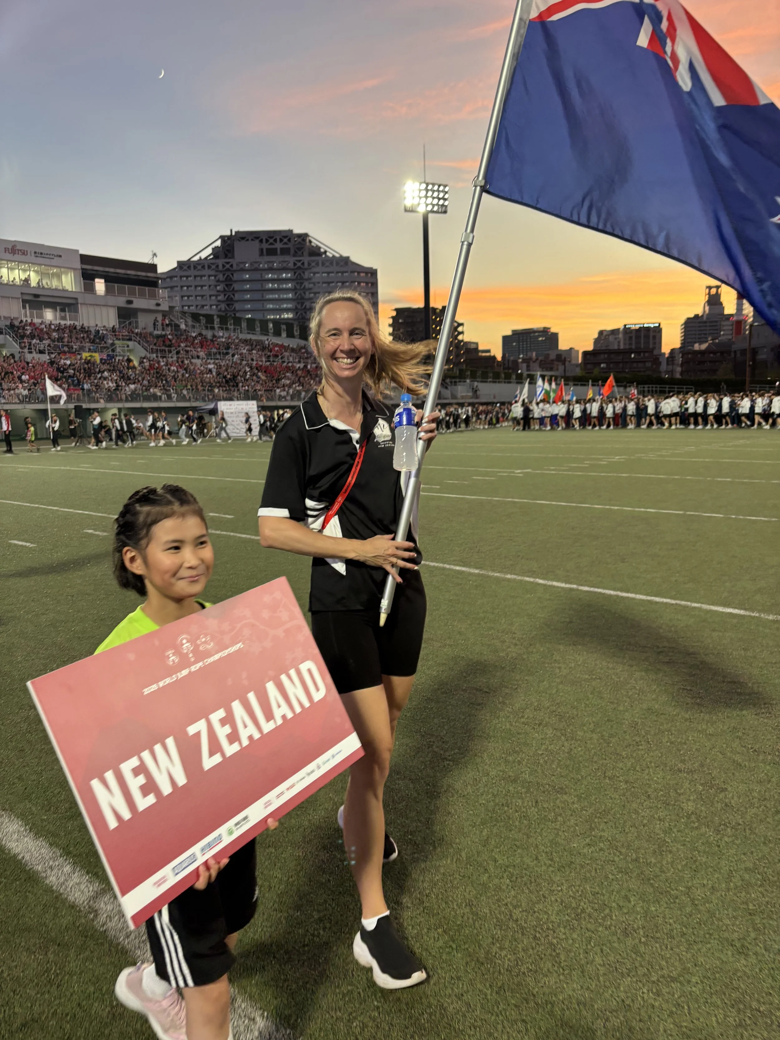 Sacha dressed in her New Zealand uniform at the World Championship's Opening Ceremony holding a New Zealand flag, a child holds a sign saying New Zealand.