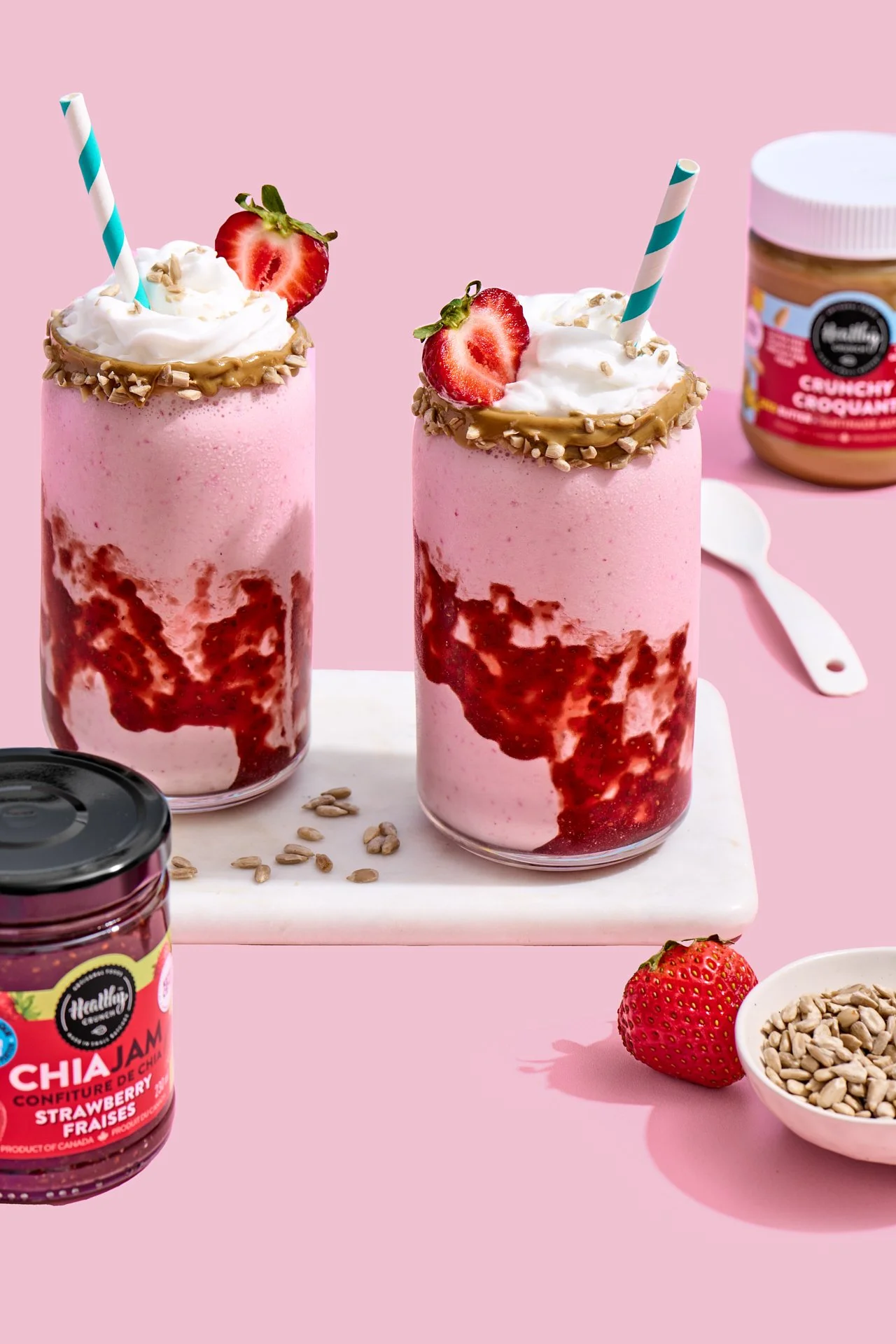 glasses of strawberry milkshakes with a strawberry sauce swirl and dollops of whipped cream on top on a pink background