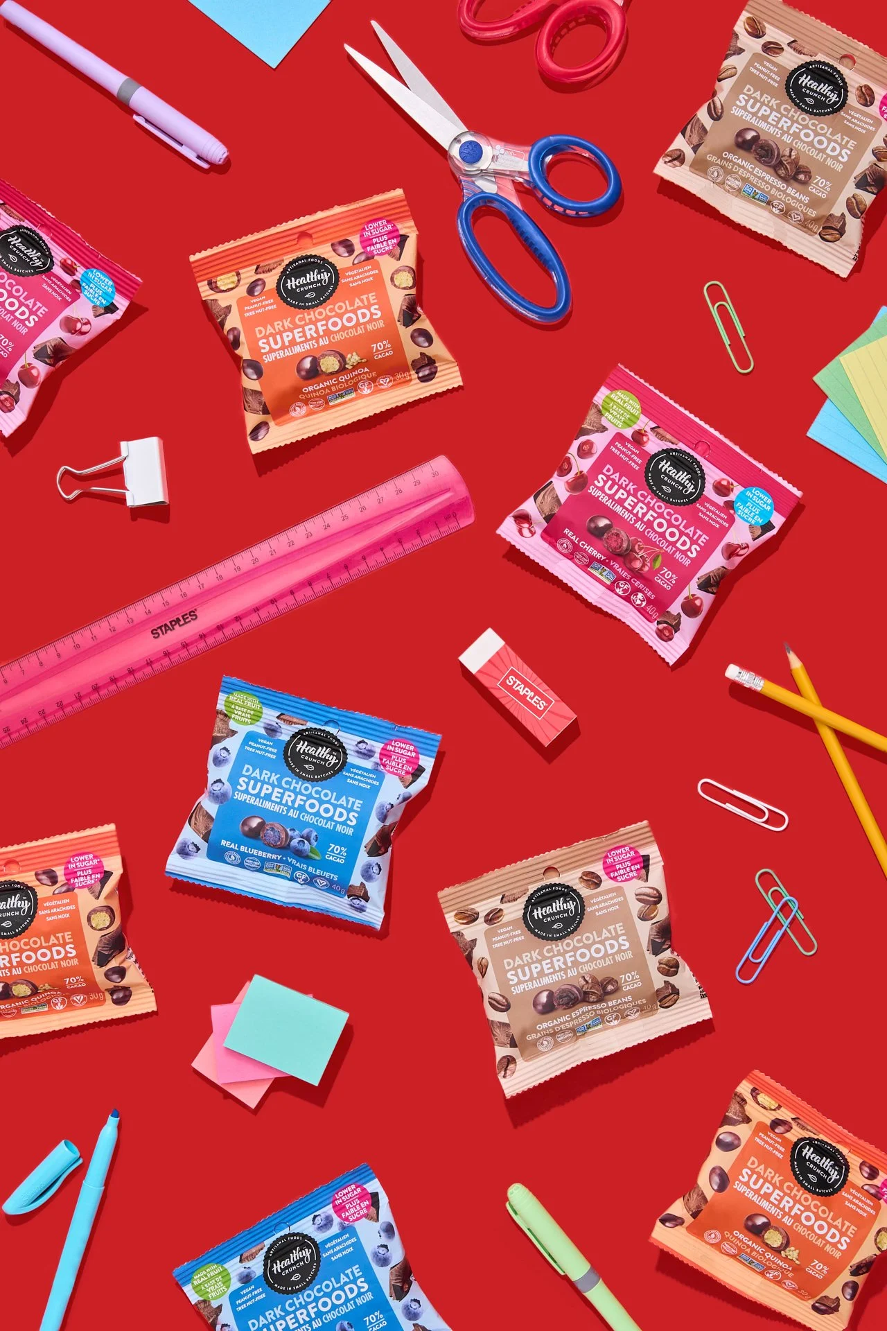 bright, colourful, vibrant, bold commercial photography with warious colorful cpg snack packages, office supplies like scissors, markers, paper clips, sticky notes, a ruler, and snack packets of organic dark chocolate superfoods spread on a red backg