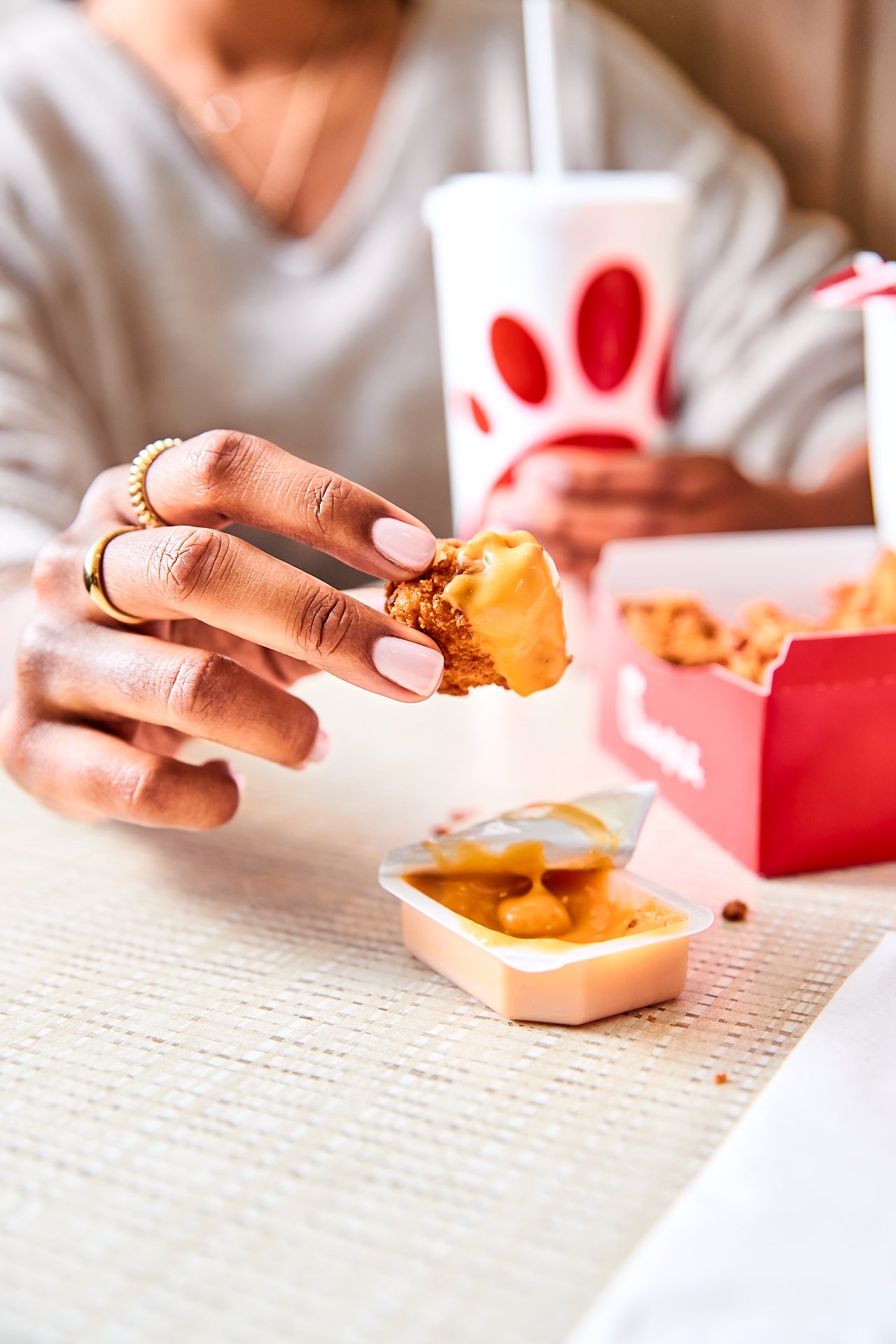 a hand holding a chick-fil-a chicken nugget dipping into chick-fil-a sauce, out of focus in background is a woman and the waffles fries and her torso