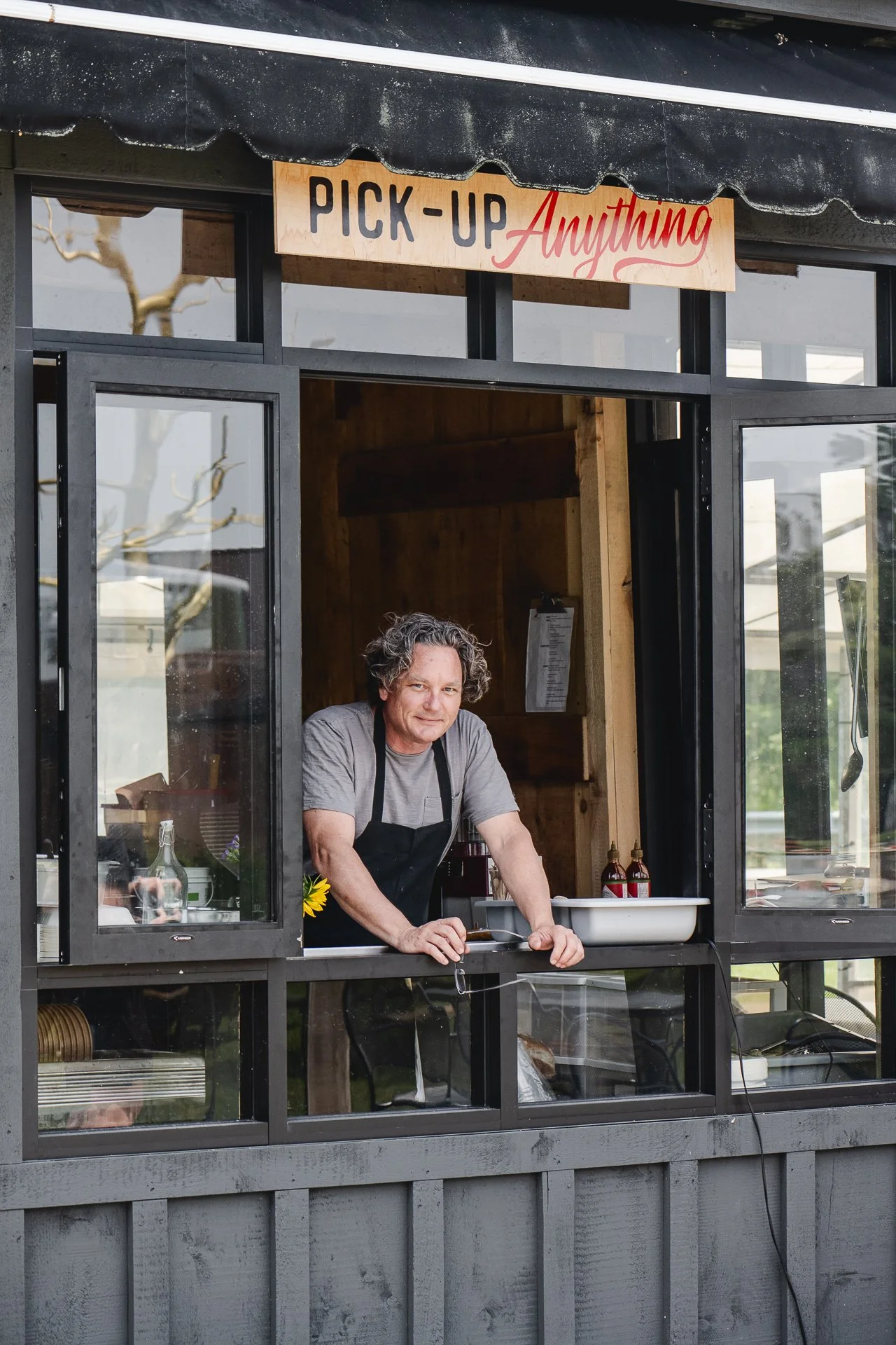 a chef leaning out the window of pick up window of a restaurant. he's wearing a black chef's apron and a warm smile on his face