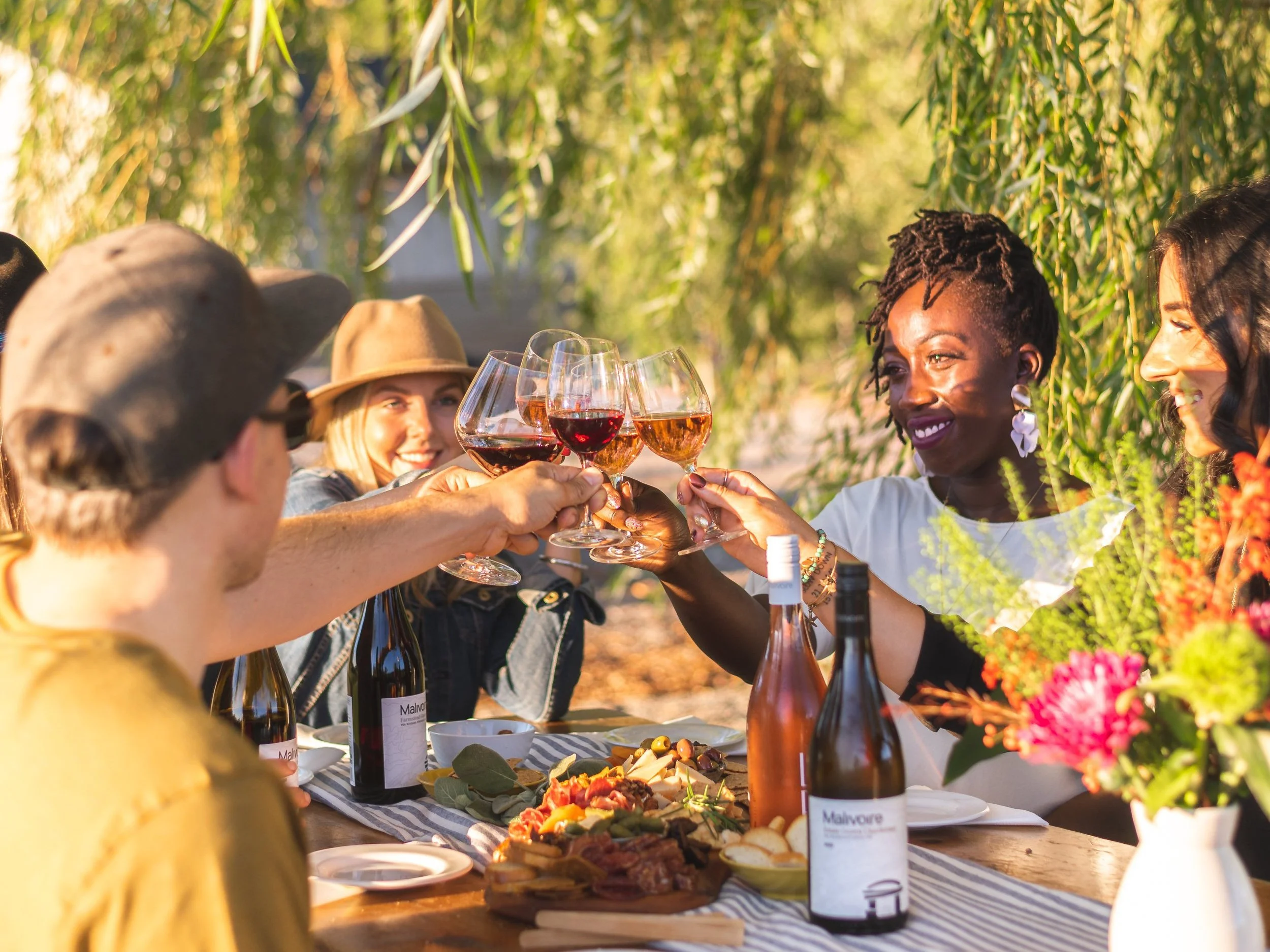 a group of friends sitting around a harvest table at golden hour at a winery cheersing glasses of red and rose wine. everyone is smilling and enjoying themselves