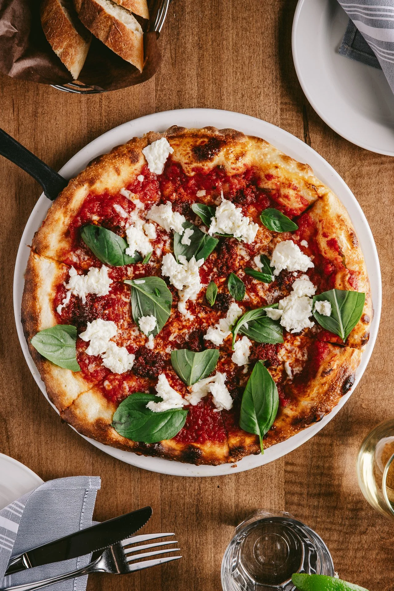 an overhead shot of a pizza on a white plate on a wooden restaurant table. pizza has tomato sauce, cheese pieces and fresh basil on top