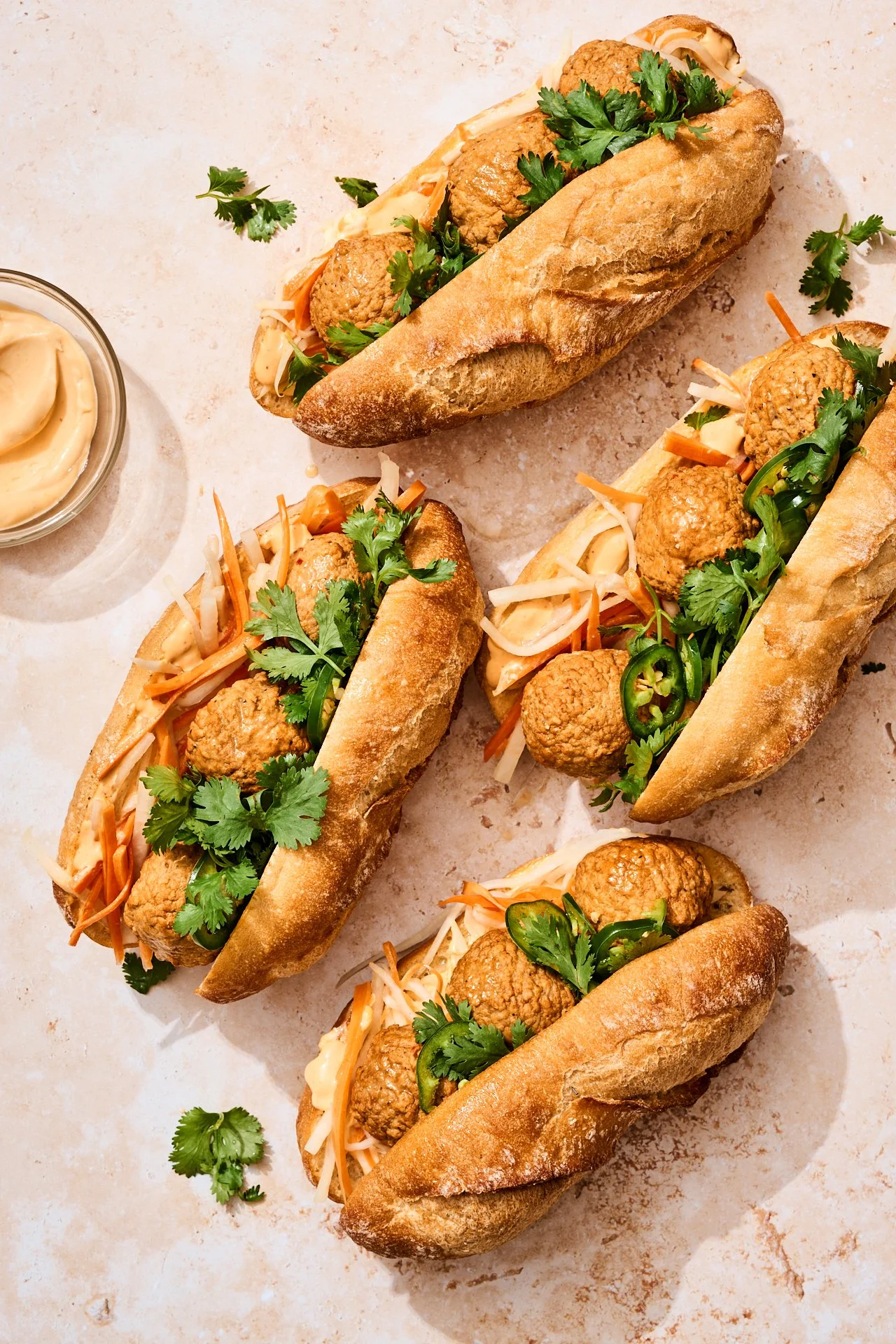 four chicken meatball subs with banh mi inspired toppings like carrot and fresh herbs and a bowl of sauce for dipping. sitting on a warm concrete backdrop