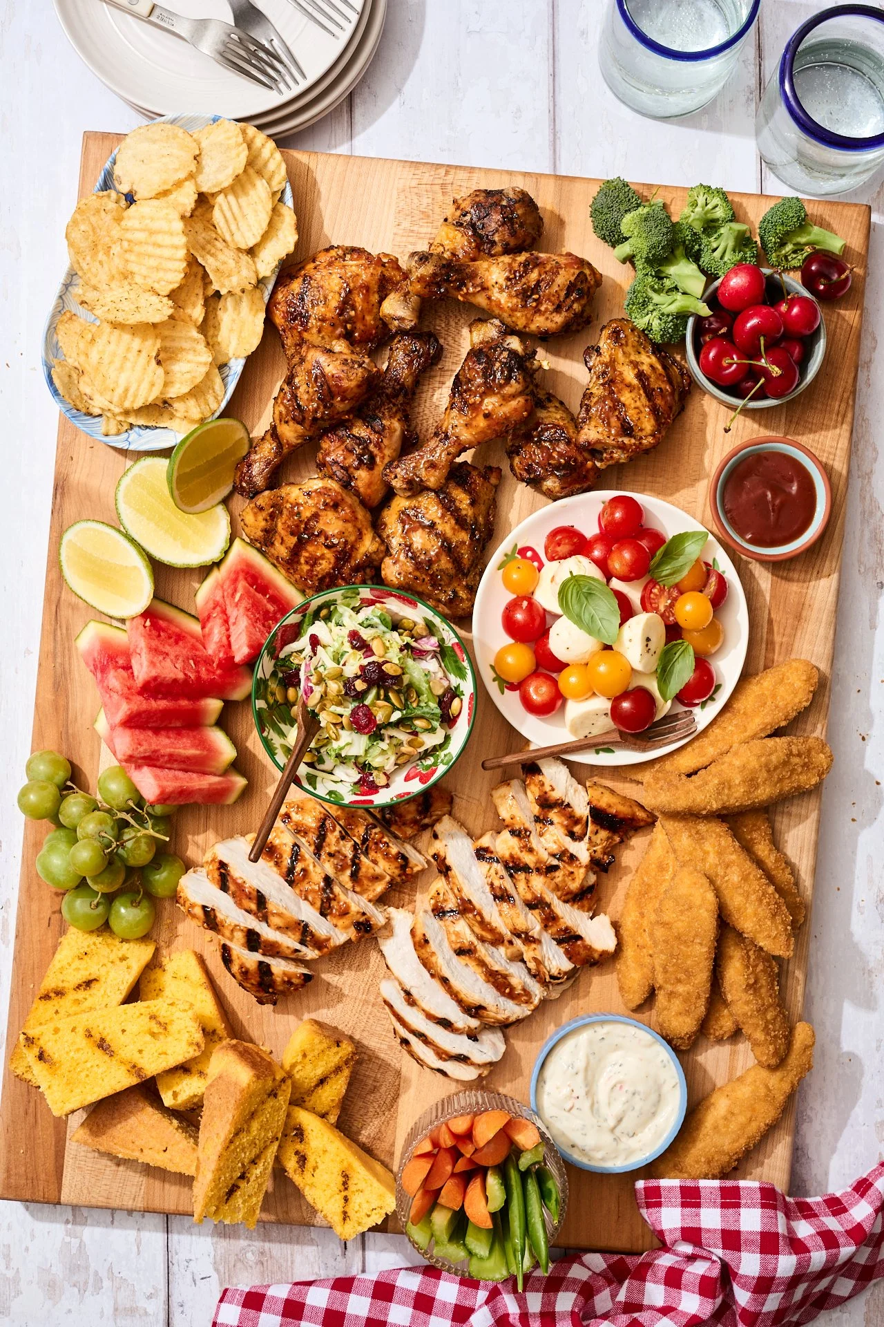 a meat board with sliced chicken breasts, chicken tenders, chicken wings, and accompanying sides like grilled cornbread, chips, tomato and cheese and basil, with a red and white napkin beside it 