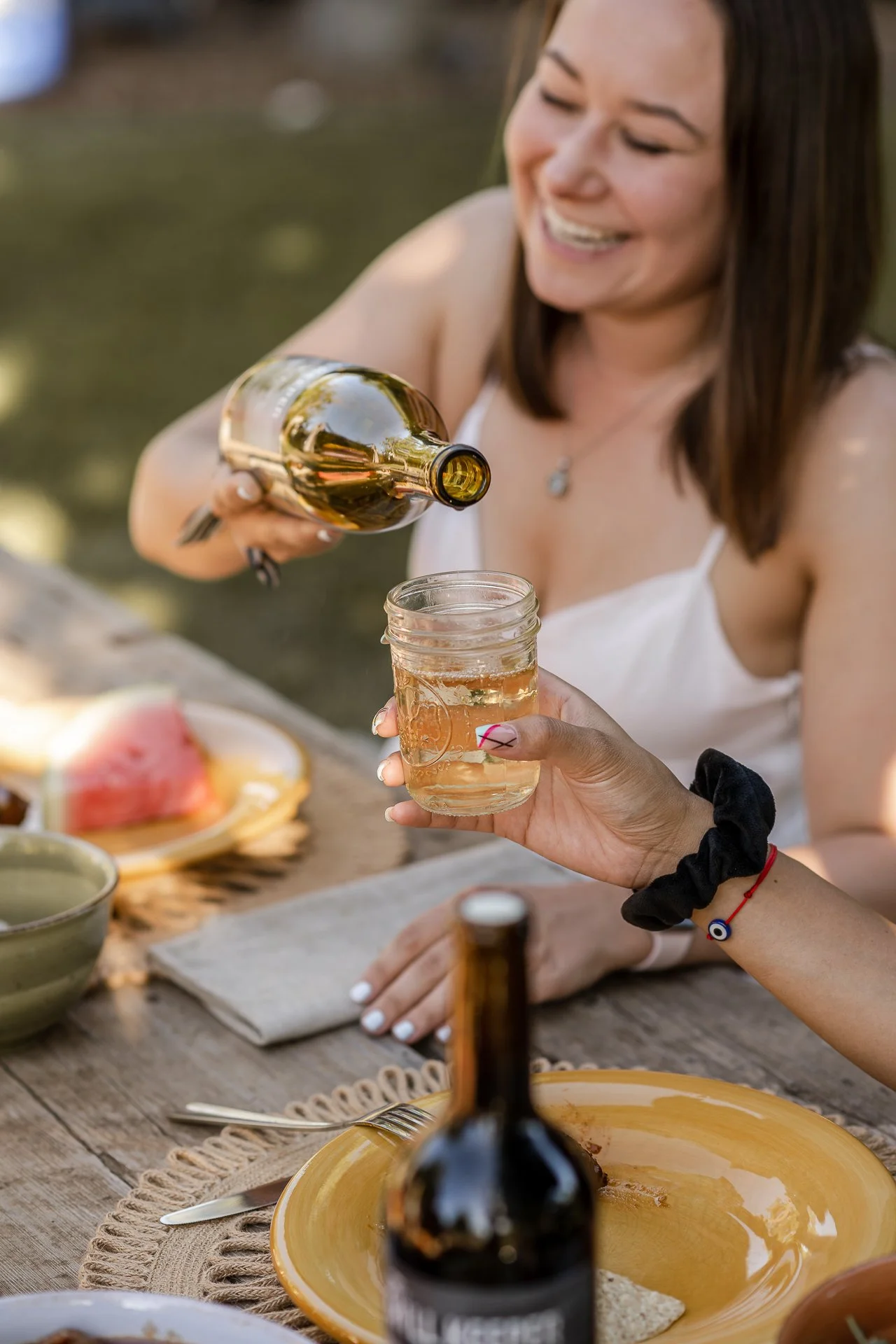 a women smiling and pouring a bottle of wine at a table with plates and summer food like watermelon