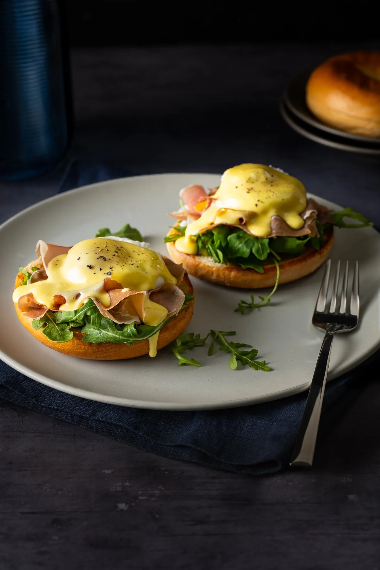 english muffin eggs benedict on a plate with arugula and hollandaise sauce oozing down against a moody dark backdrop