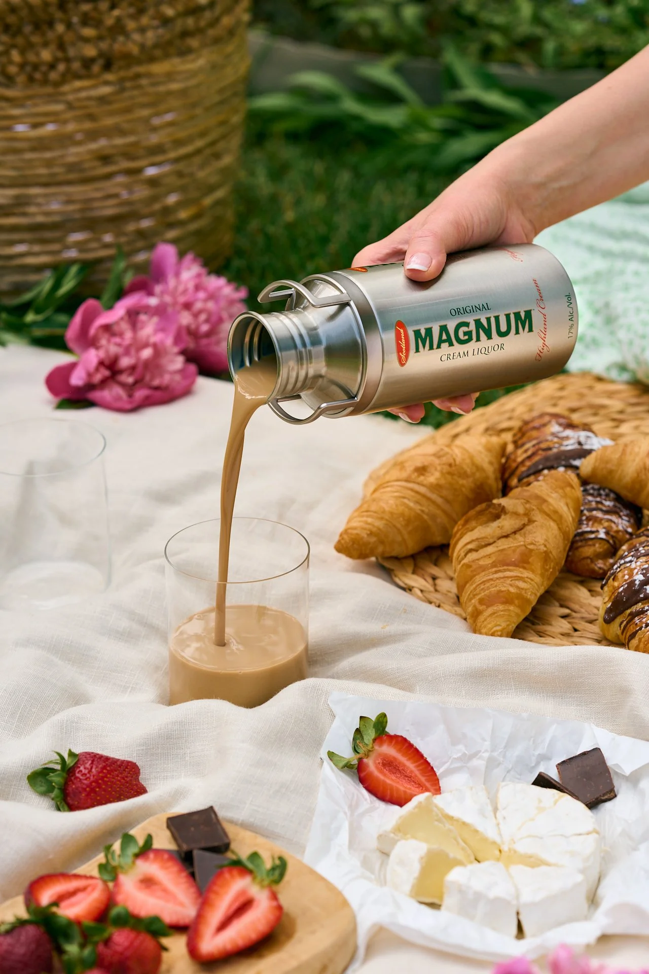 a canister of magnum cream liqueur being poured into a glass resting on a picnic blanket and surrounded with strawberries and chocolate, croissants and fresh flowers