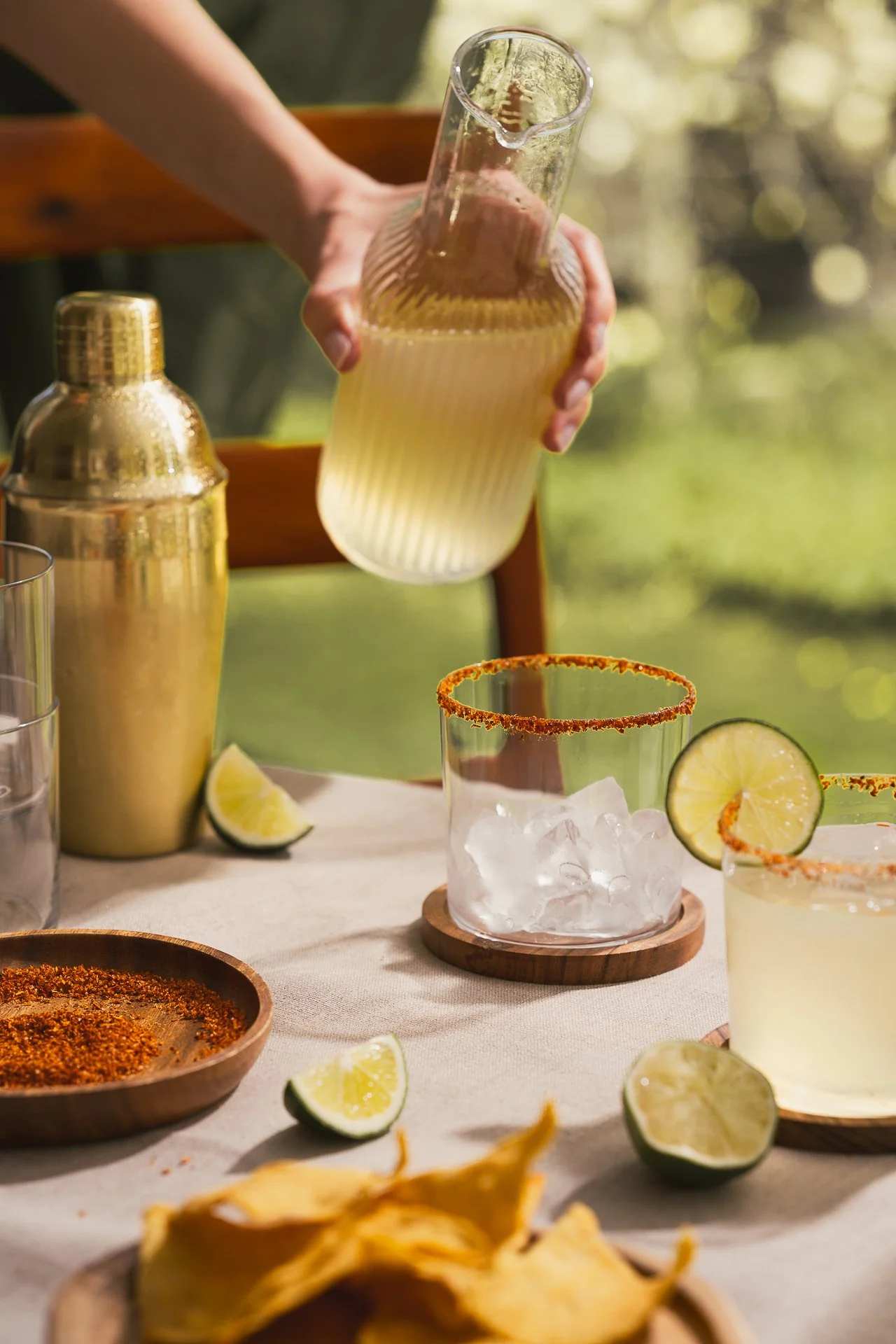 a hand pouring a yellow drink into a rimmed glass on a table surrounded with limes and tortilla chips