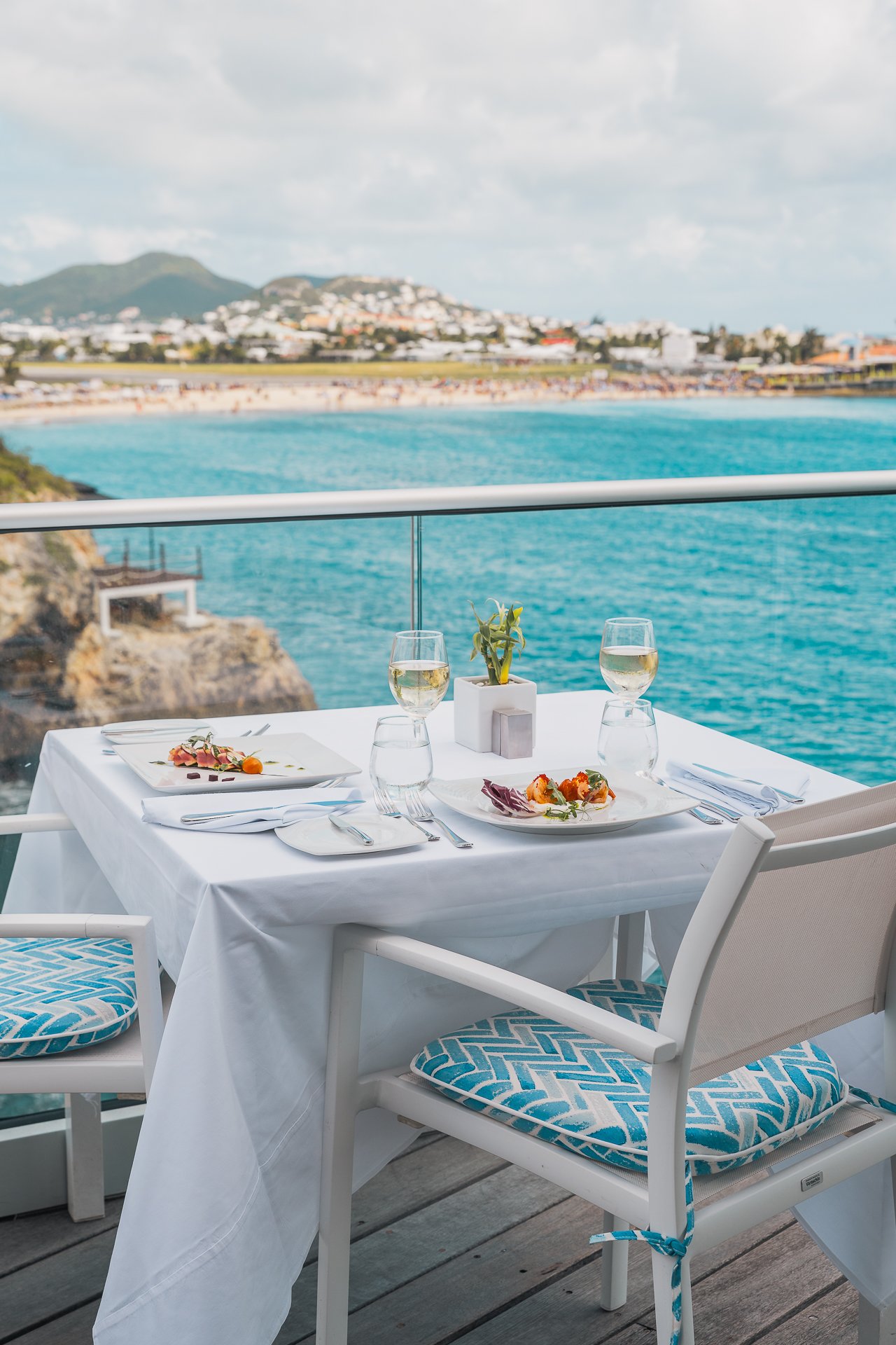 a table for two at a caribbean restaurant at the st maarten resort ocean point. the ocean, beach and mountain are seen in the distance with a white tablecloth, plates of food and glasses of wine on the table