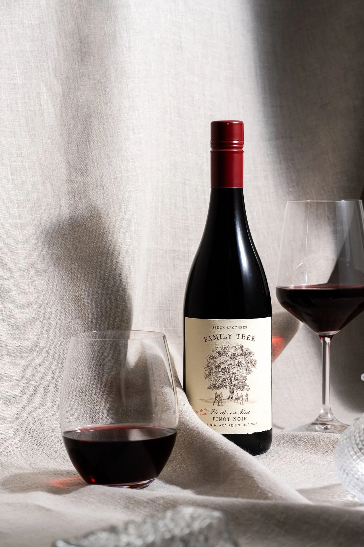 Commercial wine photography featuring a henry of pelham family tree wine bottle and two glasses styled against a neutral textured backdrop. Ontario or niagara winery photography.