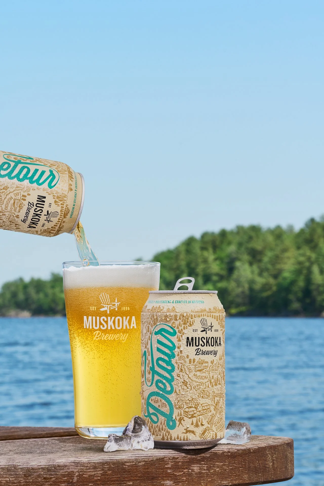 beer being poured into a glass resting on a wooden table with a can beside it and a lake in the background
