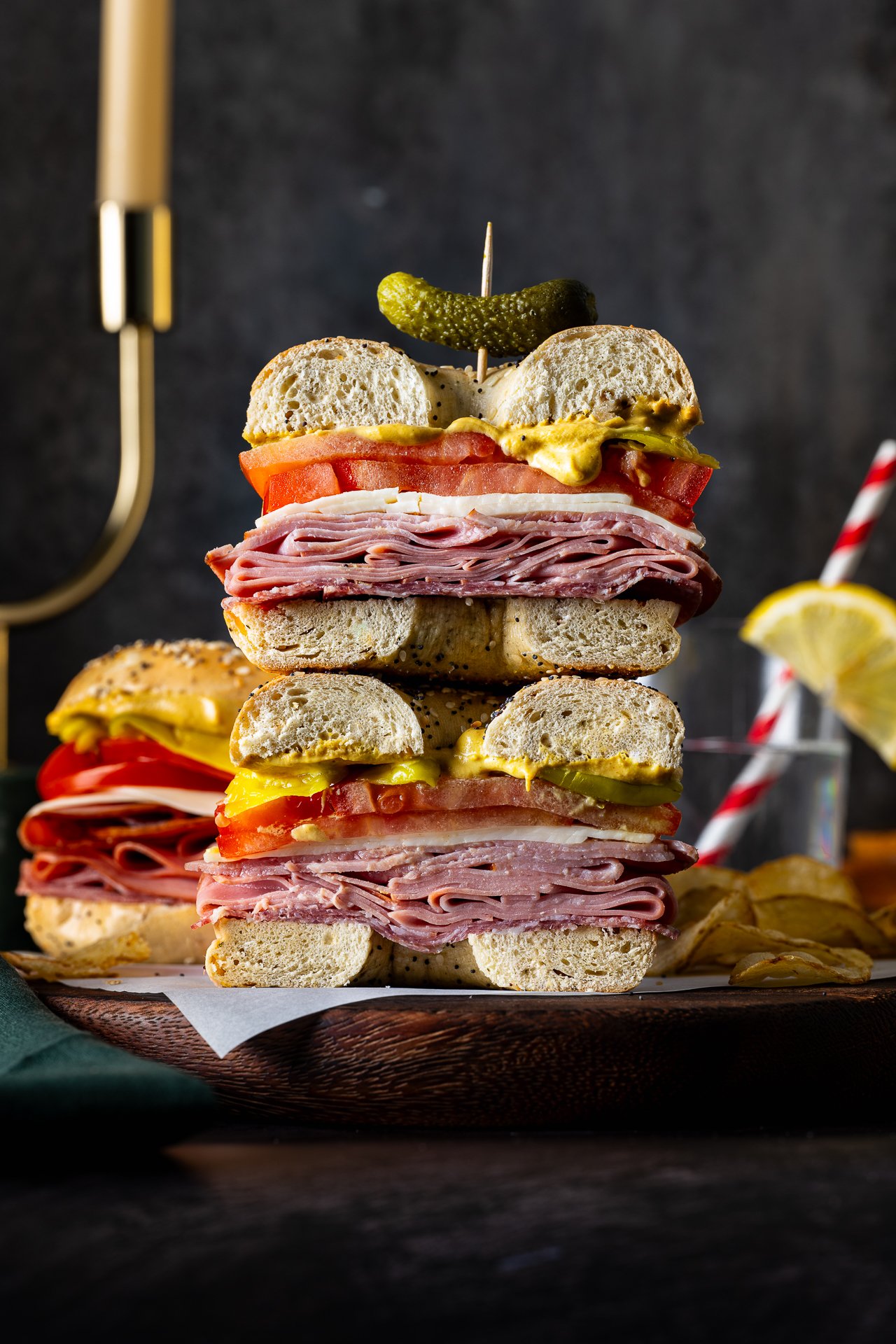 A stack of assorted deli sandwiches with meats, cheeses, tomatoes, pickles, mustard, and sandwich buns, topped with a pickle spear and toothpick.