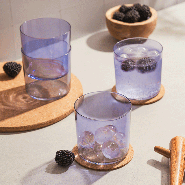 a stop motion of kilne's concord purple cups resting on cork coasters on top of a counter. a pitcher of sparkling water pours some into a glass and drops blackberries into the glass