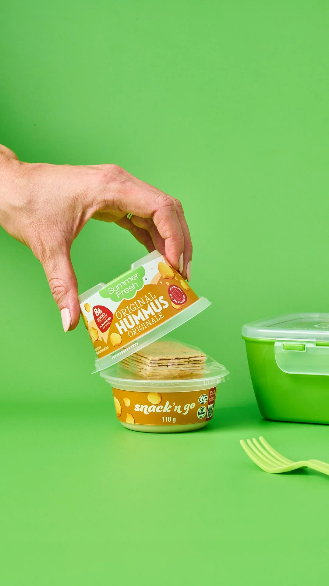 bold, colorful, vibrant lifestyle food photography of Summer Fresh's snack product of individual containers of hummus and crackers with a hand removing the lid against a bright green background.