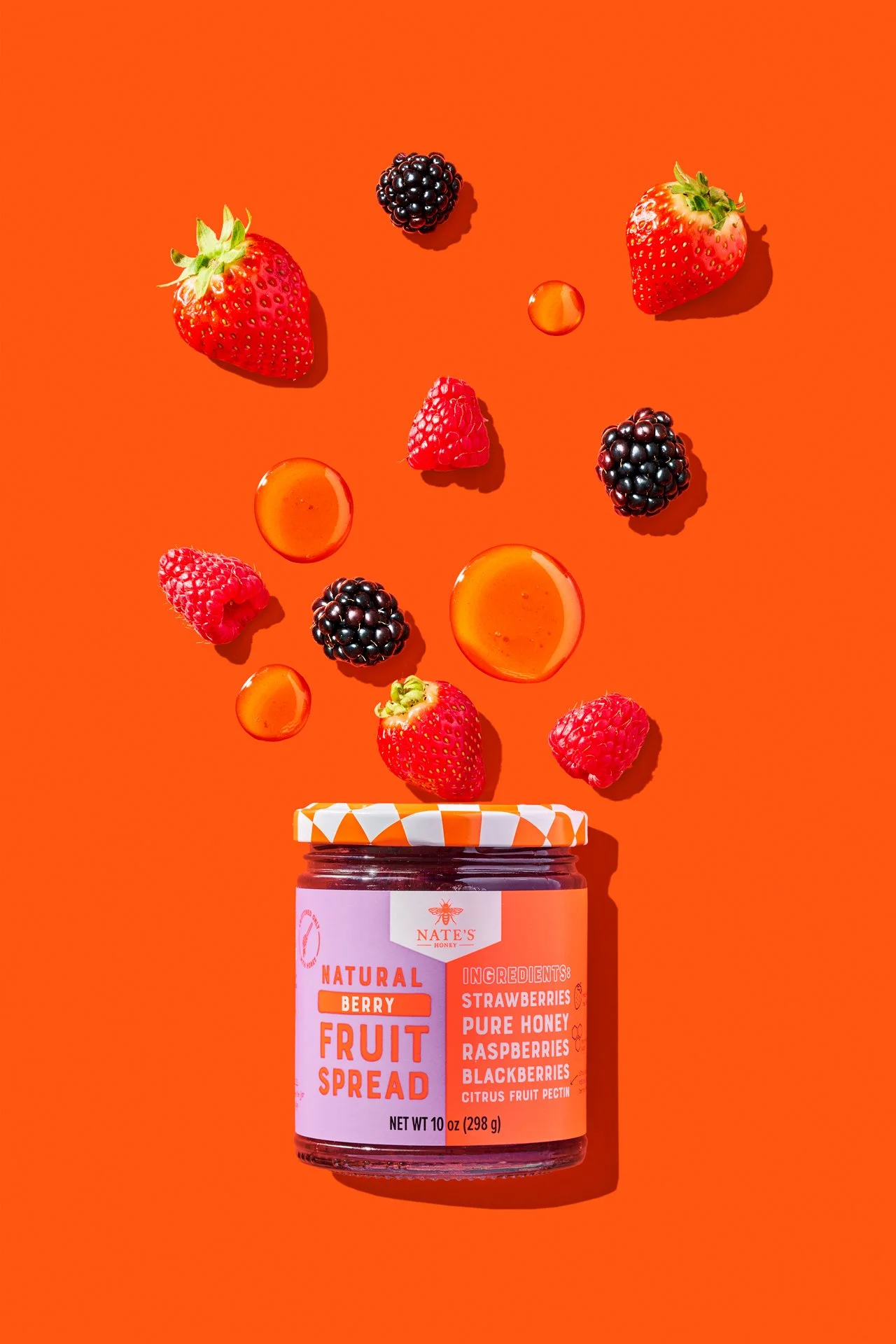 Commercial food photography of nate's honey fruit spread jar surrounded by floating berries and droplets of honey on a bright orange background.