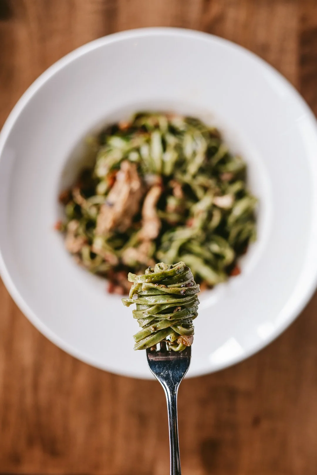 Close-up of a fork with cooked spaghetti over top of a blurred bowl of spaghetti on a wooden surface.