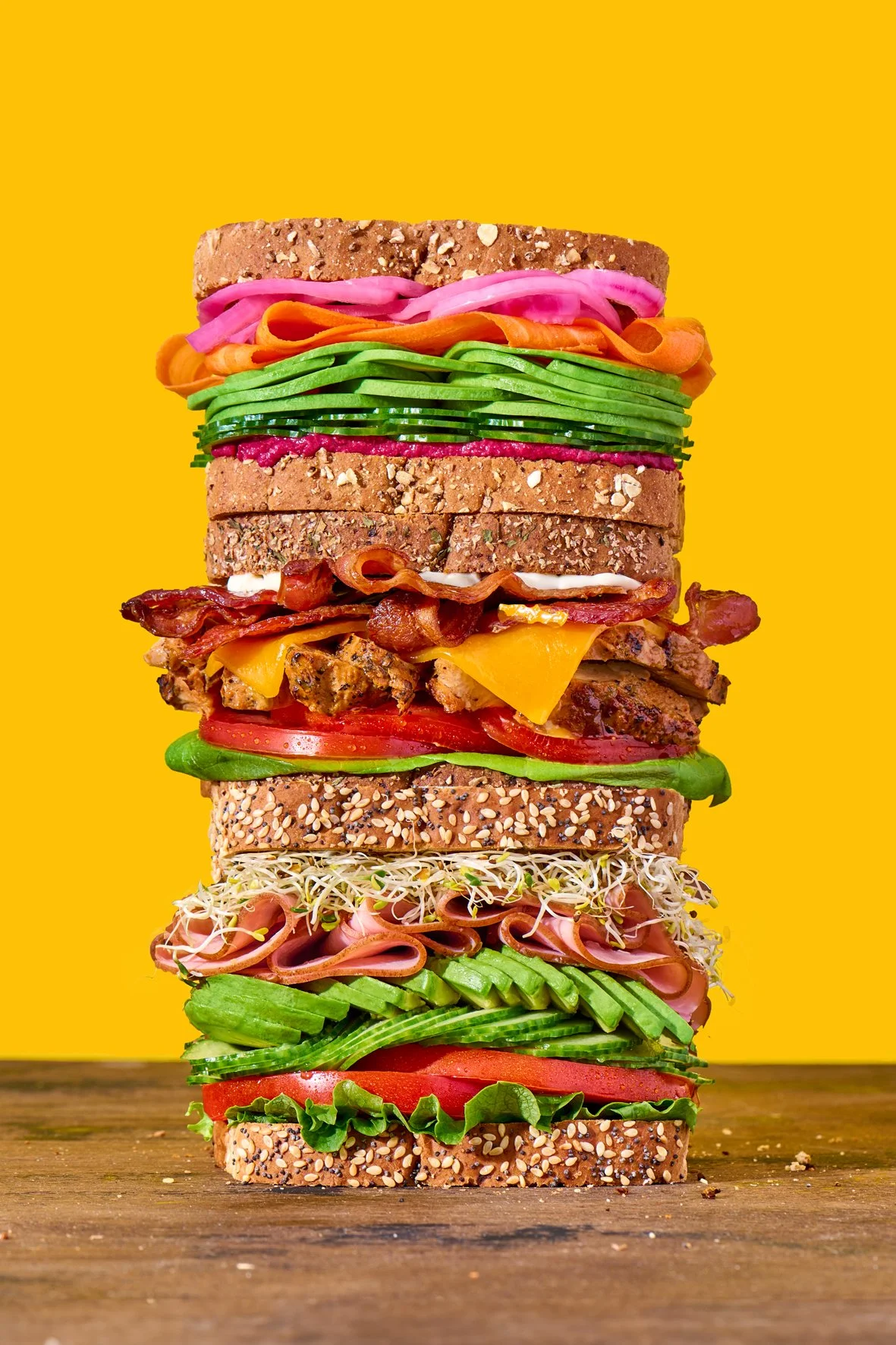 food styled sandwich stack of three sandwiches with fresh veggies, bacon, sliced meat on a wooden surface