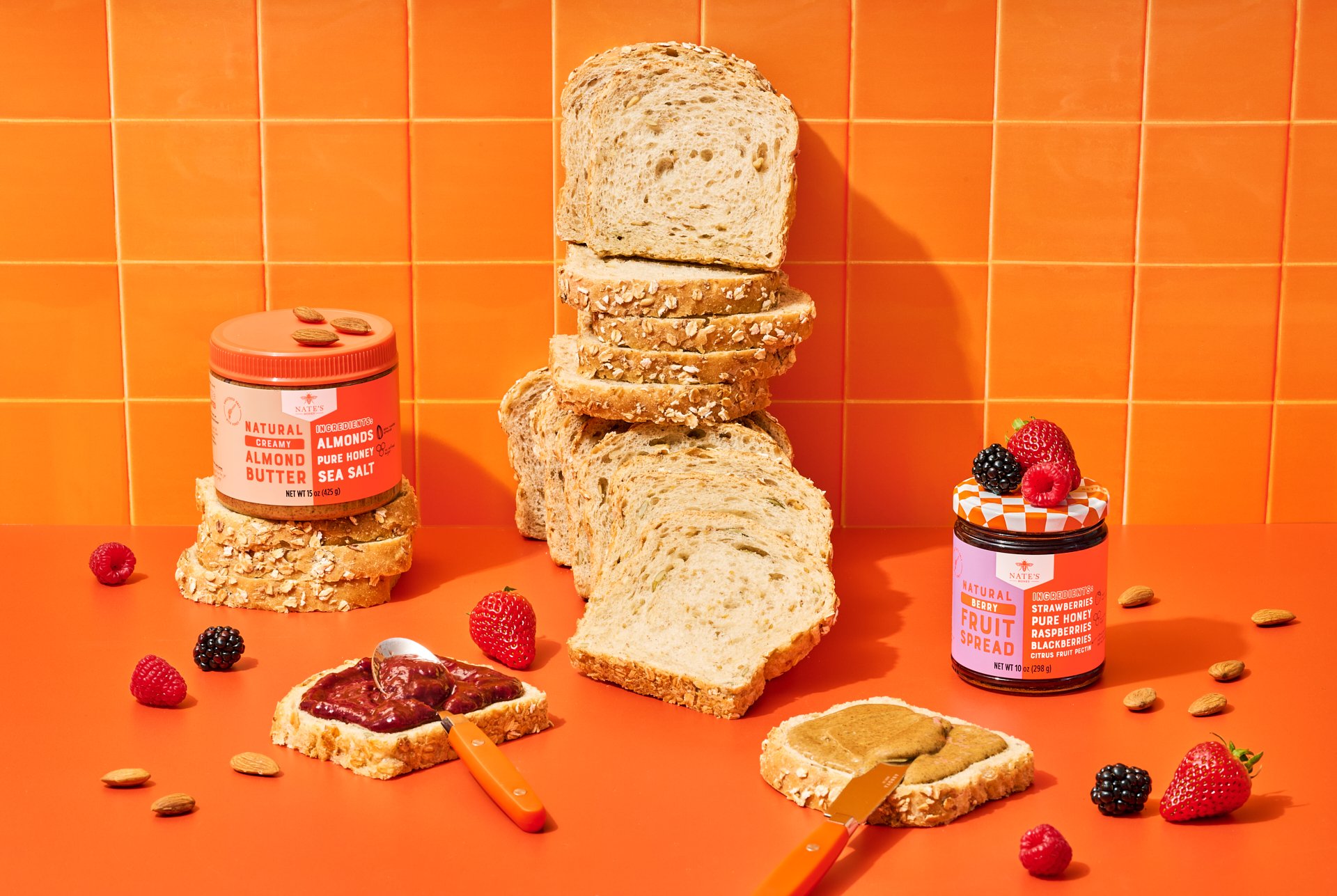 studio product photograph with a stack of bread, nate's peanut butter and mixed berry fruit spread on pieces of bread on an orange background