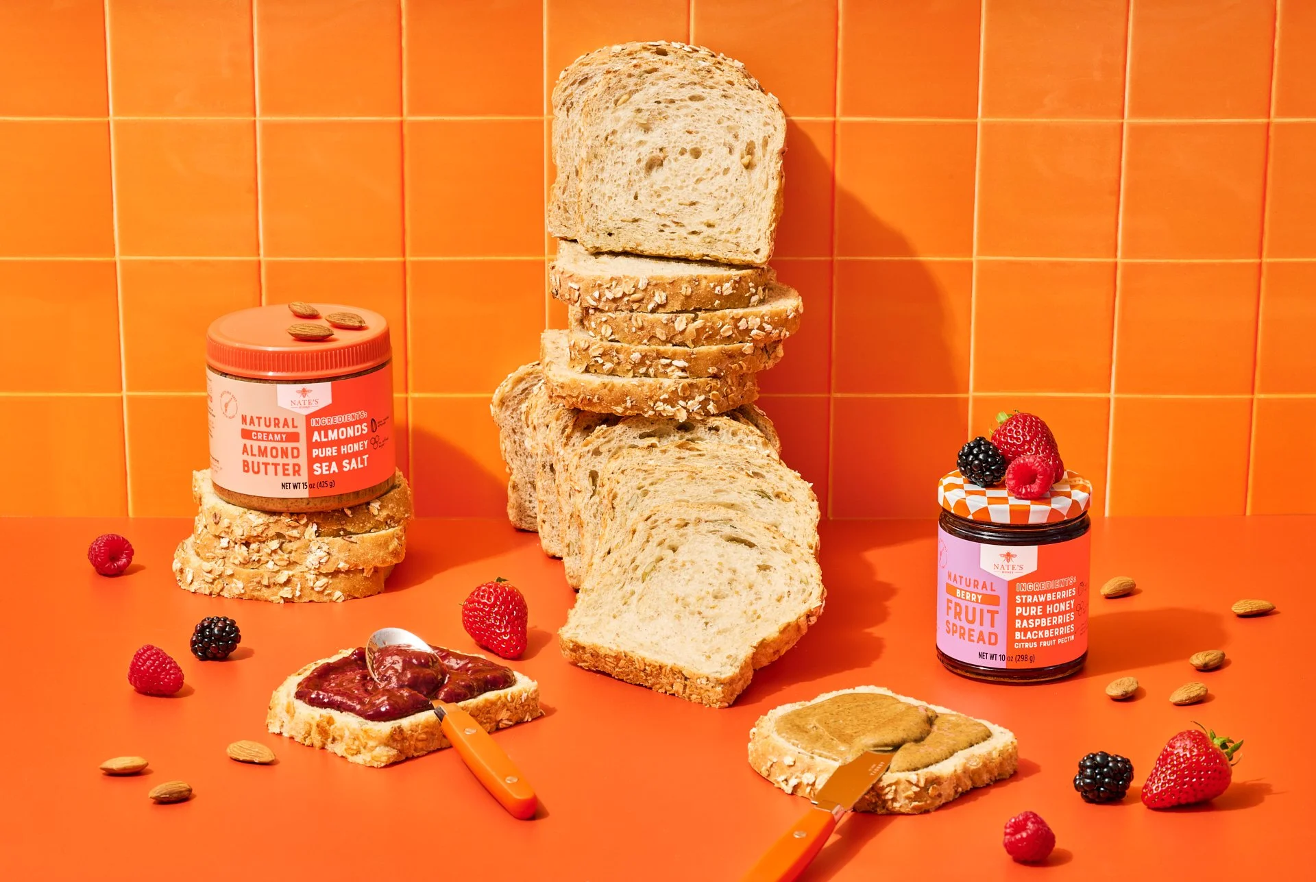studio product photograph with a stack of bread, nate's peanut butter and mixed berry fruit spread on pieces of bread on an orange background