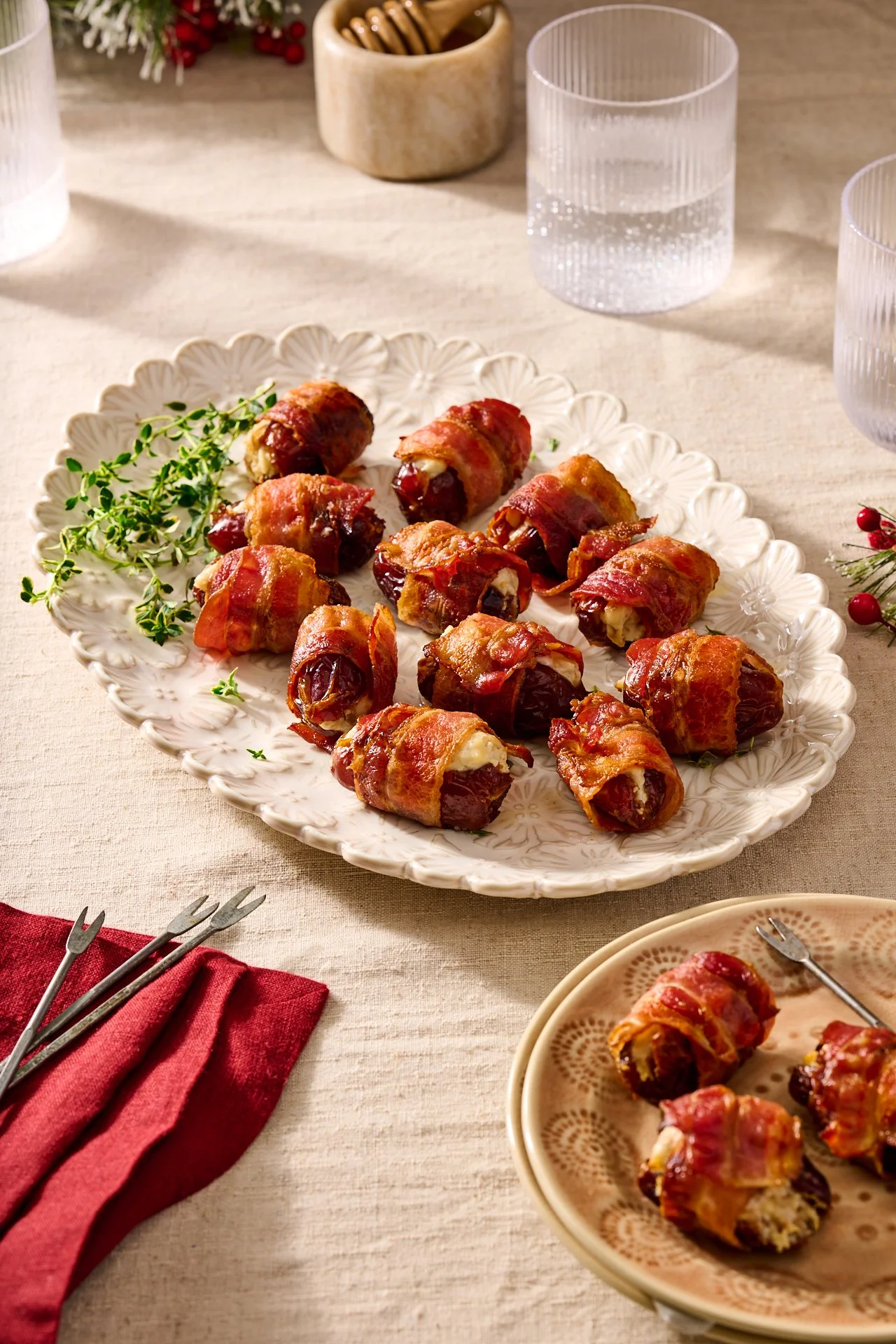 a platter in the centre of the image with bacon wrapped dates stuffed with goat cheese and walnuts and drizzled with honey and sprinkled with time, set on a pale linen tablecloth