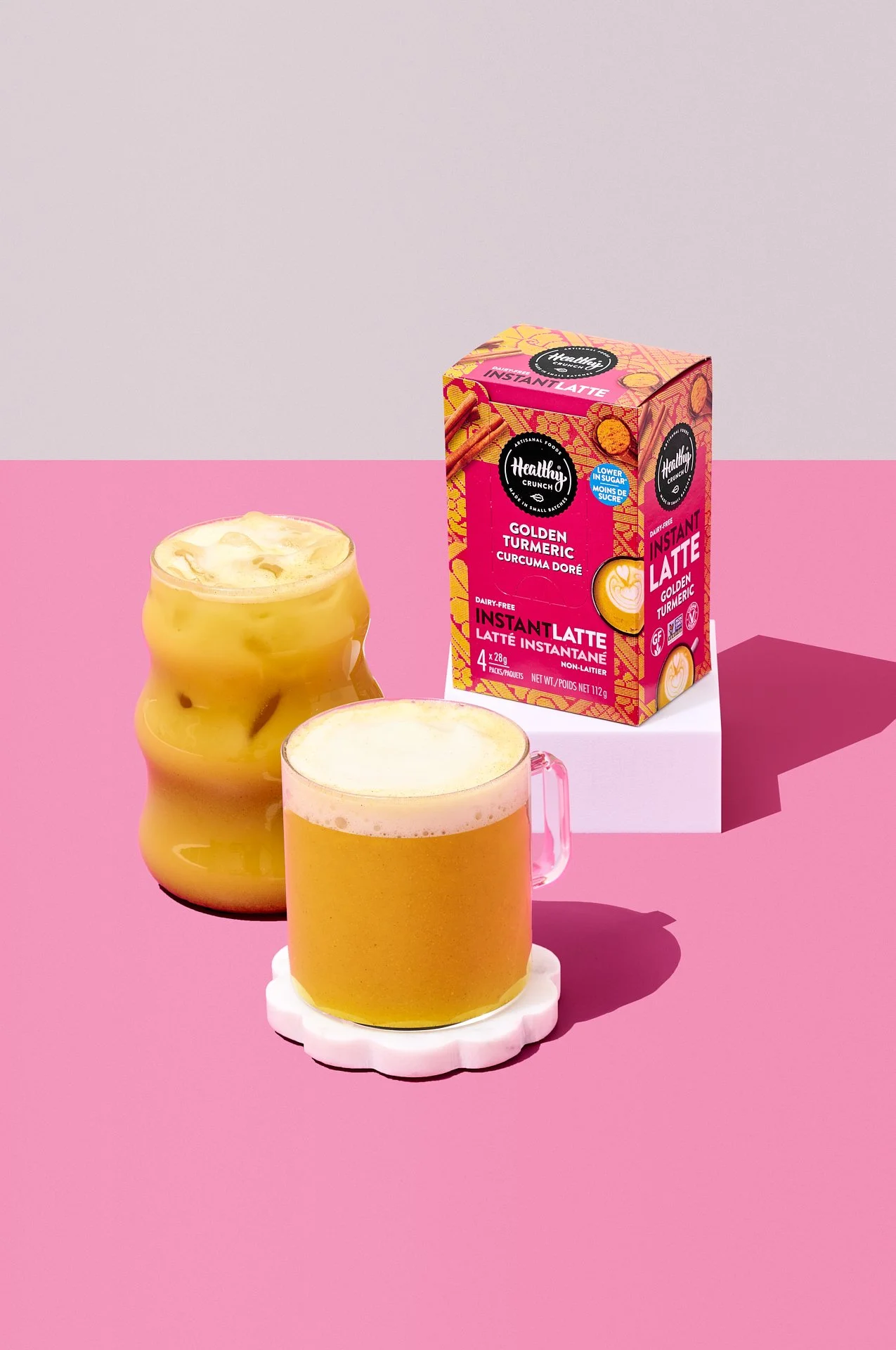 bold, colorful, vibrant commercial beverage photograph in a production studio of healthy crunch turmeric/golden latte mix with a hot and cold prepared drink styled on a bright pink set.
